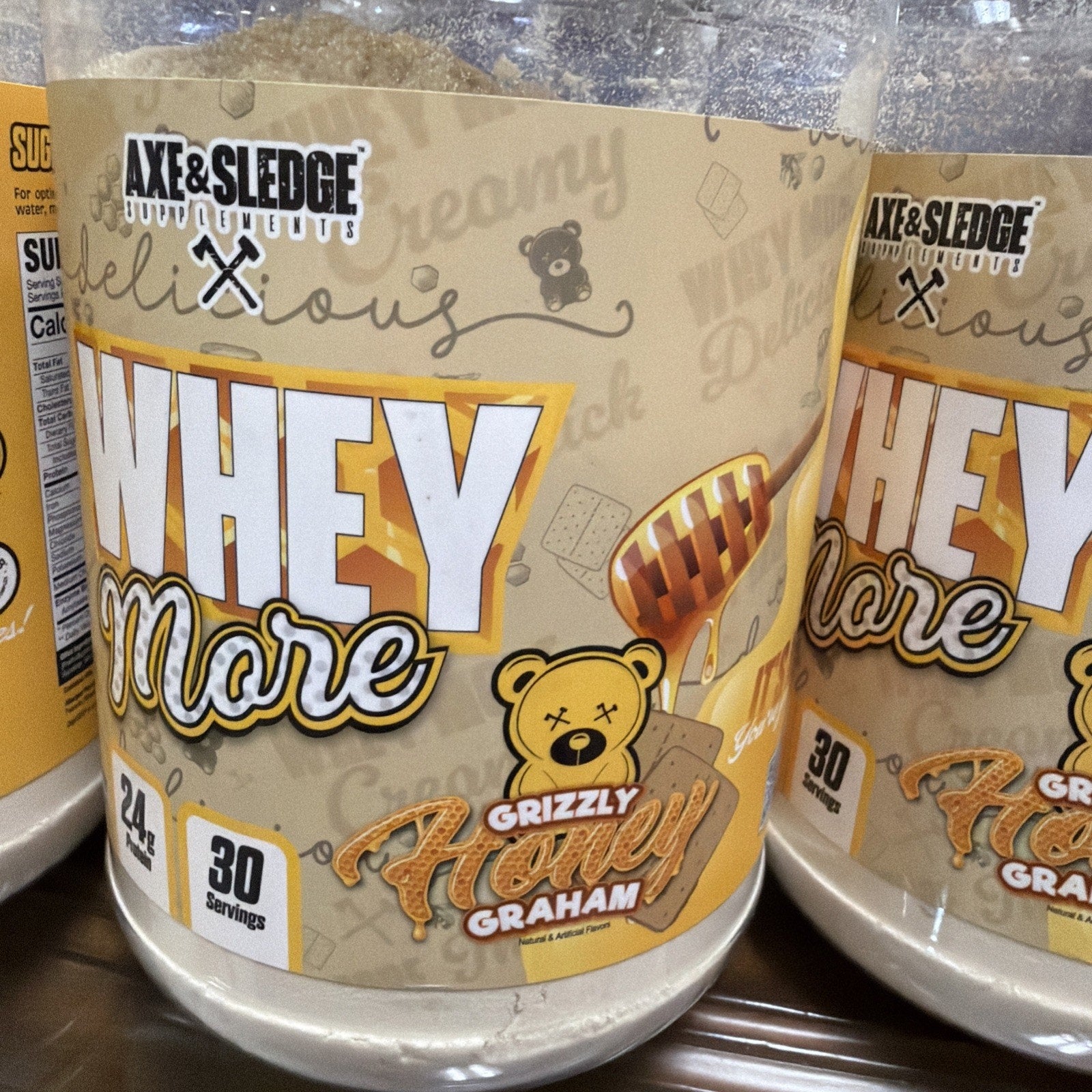 Clear plastic container of Axe & Sledge Whey More protein powder in Grizzly Honey Graham flavor, with label highlighting 24 grams of protein and 30 servings per container, featuring a honey dipper and teddy bear illustration.