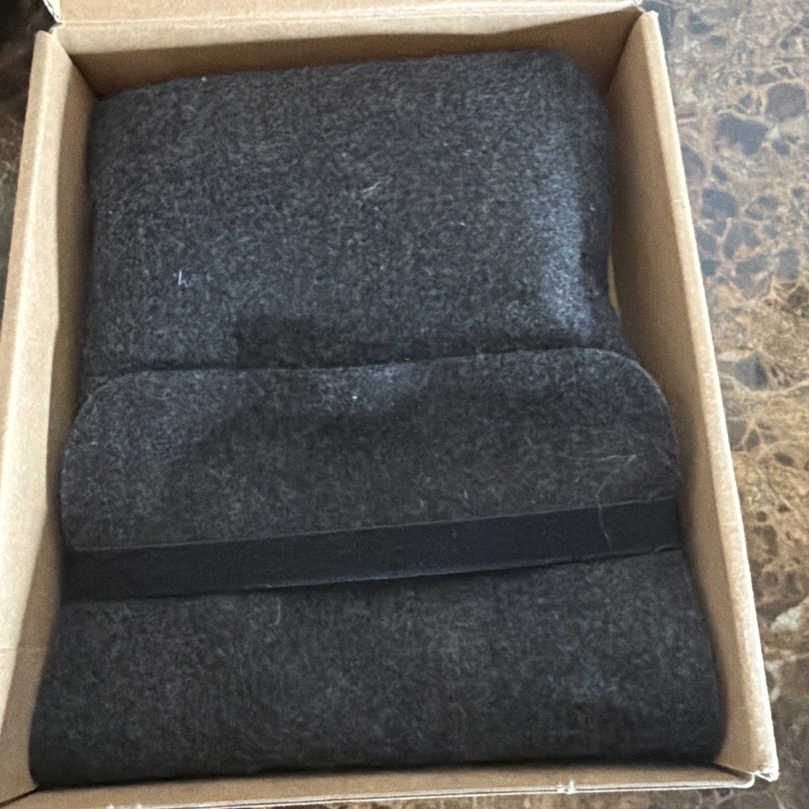 Dark gray fleece blanket neatly folded inside a plain cardboard box with a black fabric strap, displayed on a marble surface.