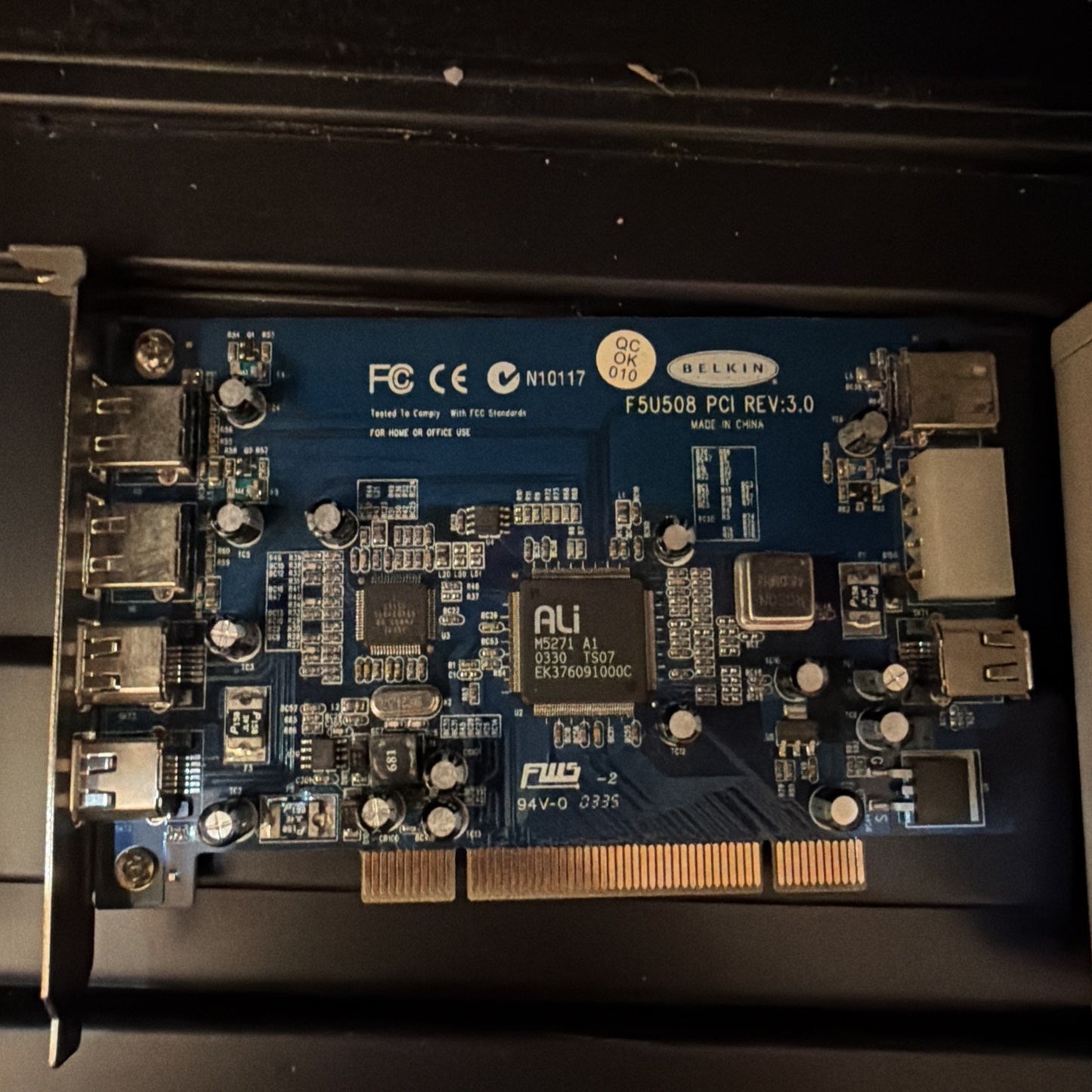 Close-up of a blue circuit board with multiple capacitors, chips, USB ports, and a PCI connector, labeled with markings including "BELKIN," certification logos, and model information.
