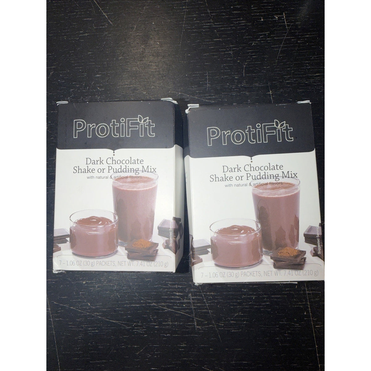 Two boxes of ProtiFit Dark Chocolate Shake or Pudding Mix, each showing a glass and bowl filled with chocolate shake or pudding alongside chocolate pieces on a dark surface.