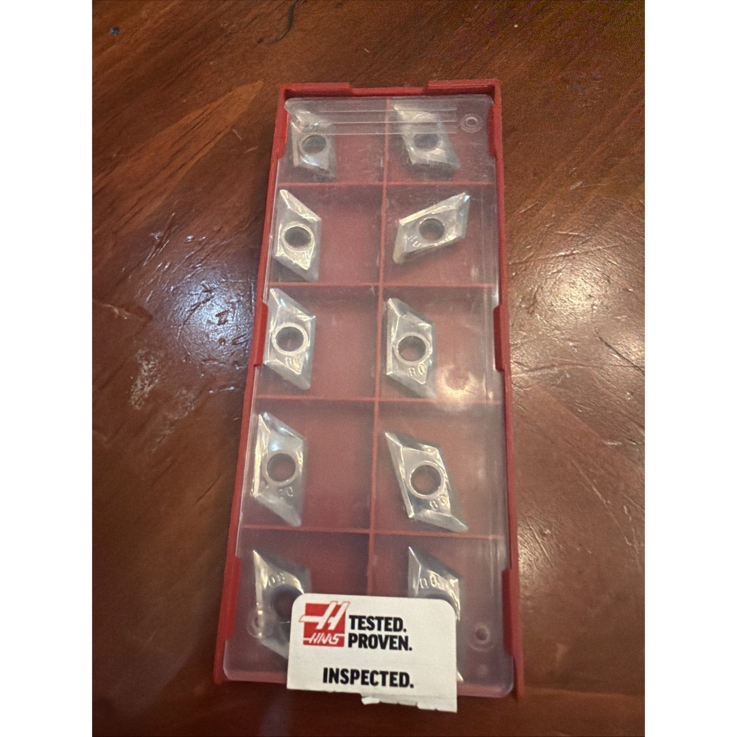 Red plastic organizer with a clear lid containing eight small silver triangular metal inserts secured inside individual compartments, resting on a wooden surface with a Haas Tested Proven Inspected label.