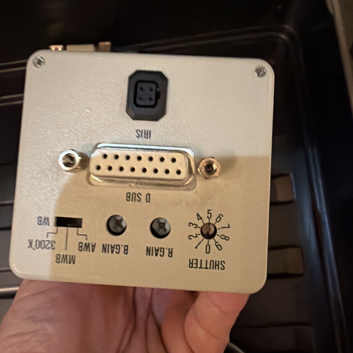 Beige-colored electronic device panel held in a hand featuring a 15-pin D-sub connector, a black 4-pin mini-DIN port labeled "MINS," two small tuning knobs marked "R.GAIN" and "B.GAIN," a switch labeled with "MWB," "3200K," and "WWB" options, and a rotary switch numbered 1 to 8 labeled "SHUTTER.