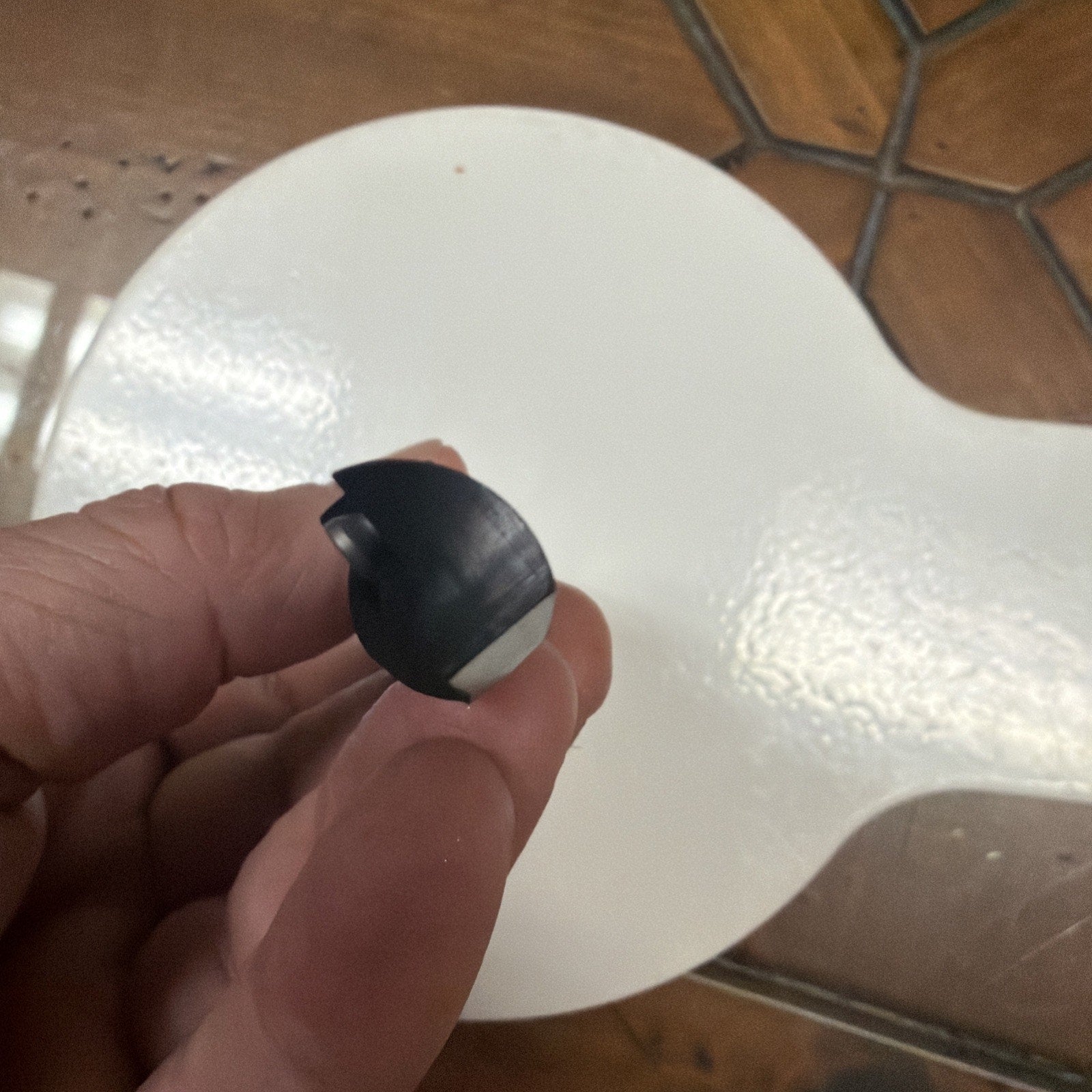 Hand holding a small black plastic or rubber component with a curved shape and a white flat section, positioned over a round white textured surface and a brown tiled floor.