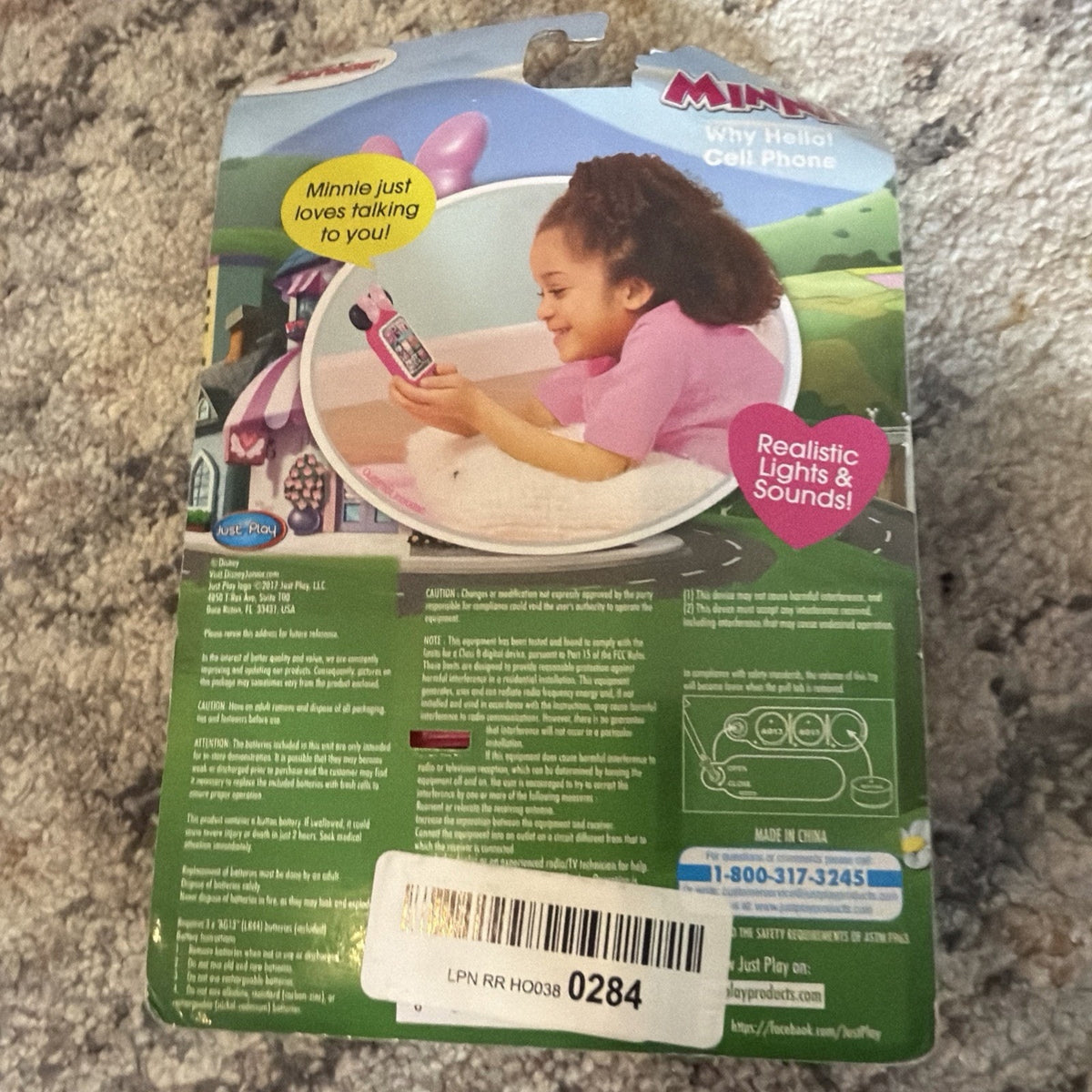 Packaging backing card for a Minnie Mouse toy phone featuring a photo of a child playing with the pink phone, with text bubbles highlighting realistic lights and sounds, placed on a carpeted surface.
