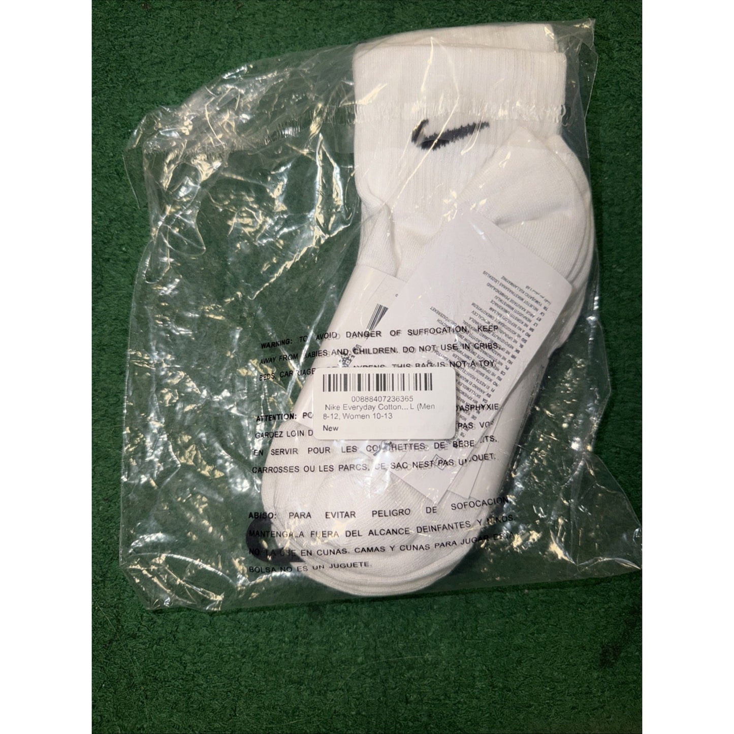 Pair of white ankle socks with black swoosh logo, packaged in clear plastic bag with safety warnings and barcode label, lying on a green fabric surface.