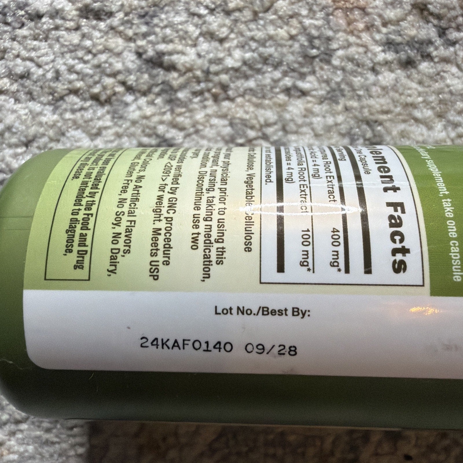 Close-up of a green supplement bottle label showing lot number 24KAF0140 and best by date 09/28, with partially visible ingredient facts and usage instructions.