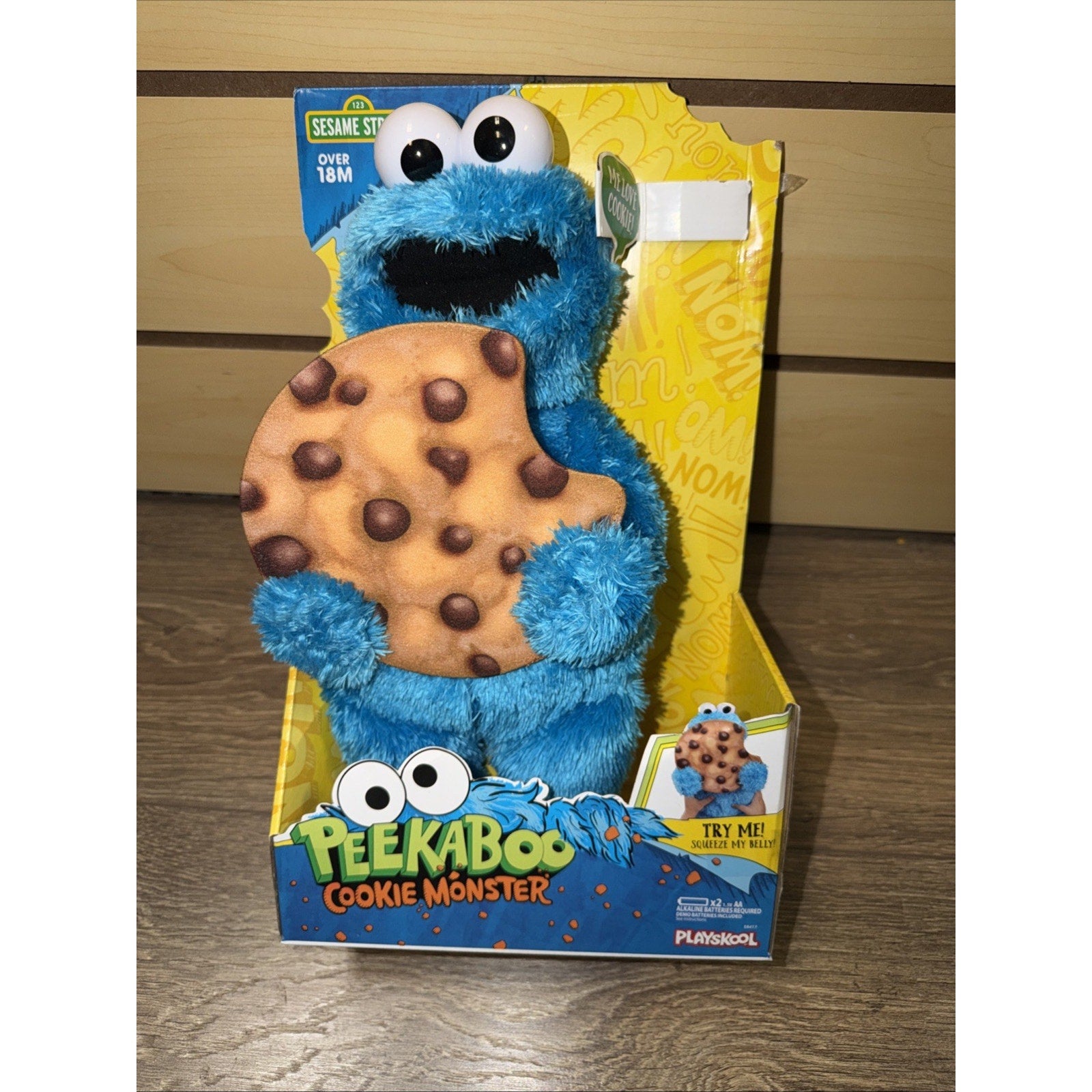 Blue fuzzy Cookie Monster plush toy holding a large bitten chocolate chip cookie, displayed in bright yellow and blue packaging with Sesame Street branding and a "Peekaboo Cookie Monster" label.
