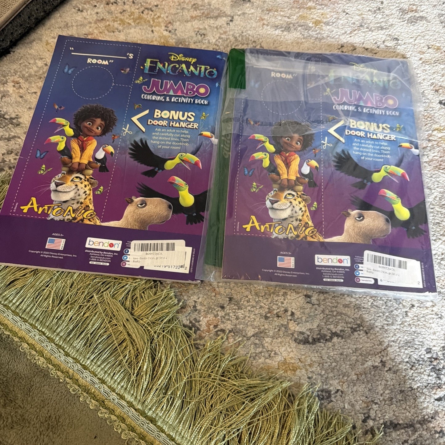 Two Disney Encanto-themed jumbo coloring and activity books featuring Antonio with animals, one unwrapped and one shrink-wrapped, resting on a carpet near a green fringed rug.