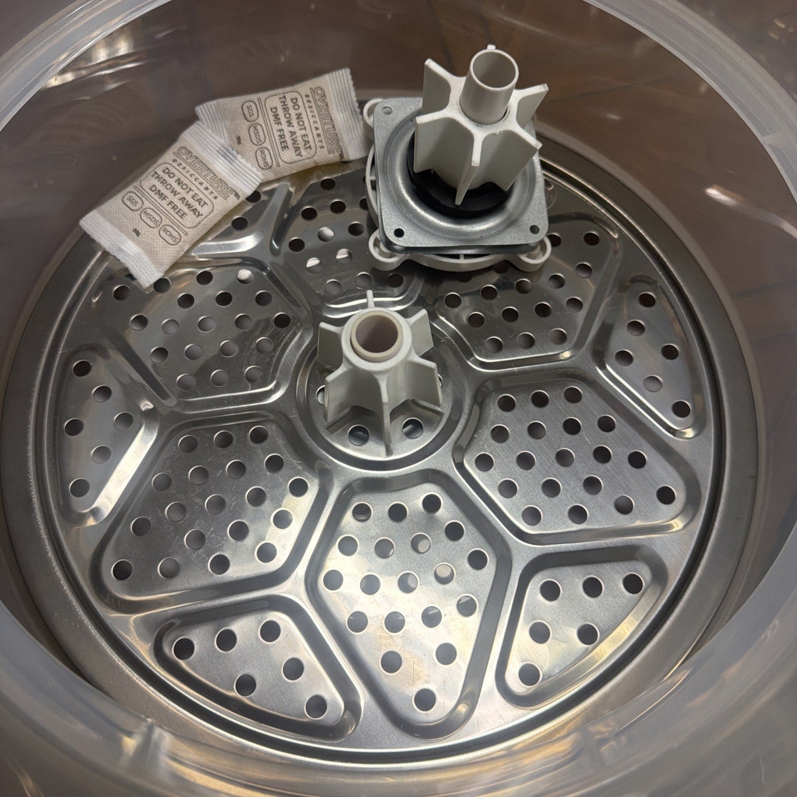 Round stainless steel perforated tray with two gray plastic mechanical parts and two small moisture absorber packets placed on top inside a clear plastic container.