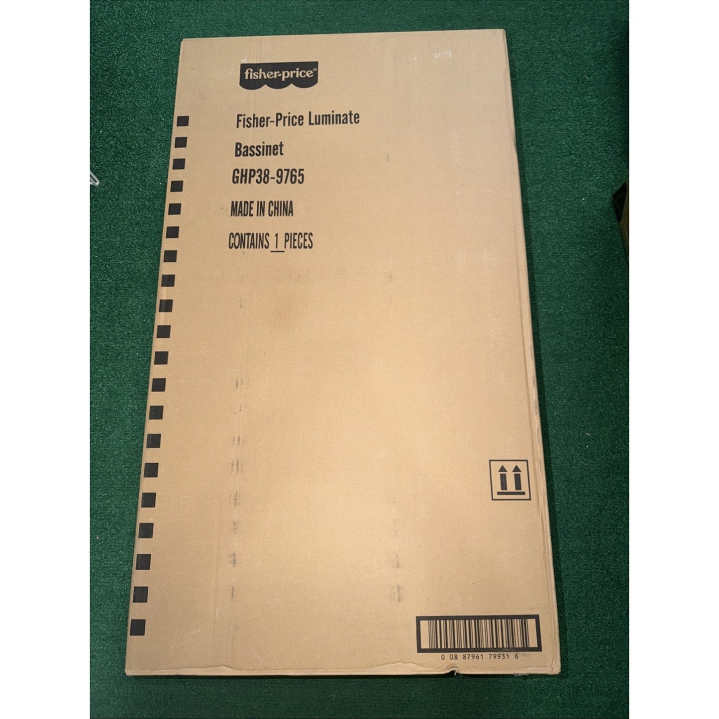 Plain cardboard shipping box for a Fisher-Price Luminate Bassinet, labeled with product name, model number, country of origin, quantity, and handling symbols on a green carpet background.