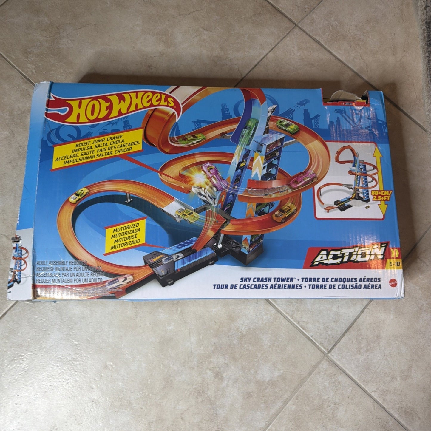 Blue Hot Wheels toy track packaging featuring an orange looped racetrack with motorized launch and spiral tower, designed for ages 5 and up, displayed on a tiled floor.