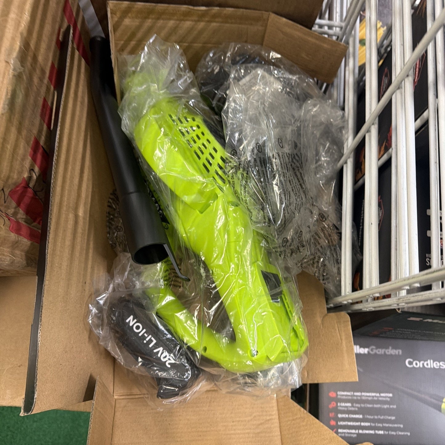 Bright green cordless handheld leaf blower with black detachable blower tube and 20V lithium battery, all wrapped in clear plastic inside a cardboard box.
