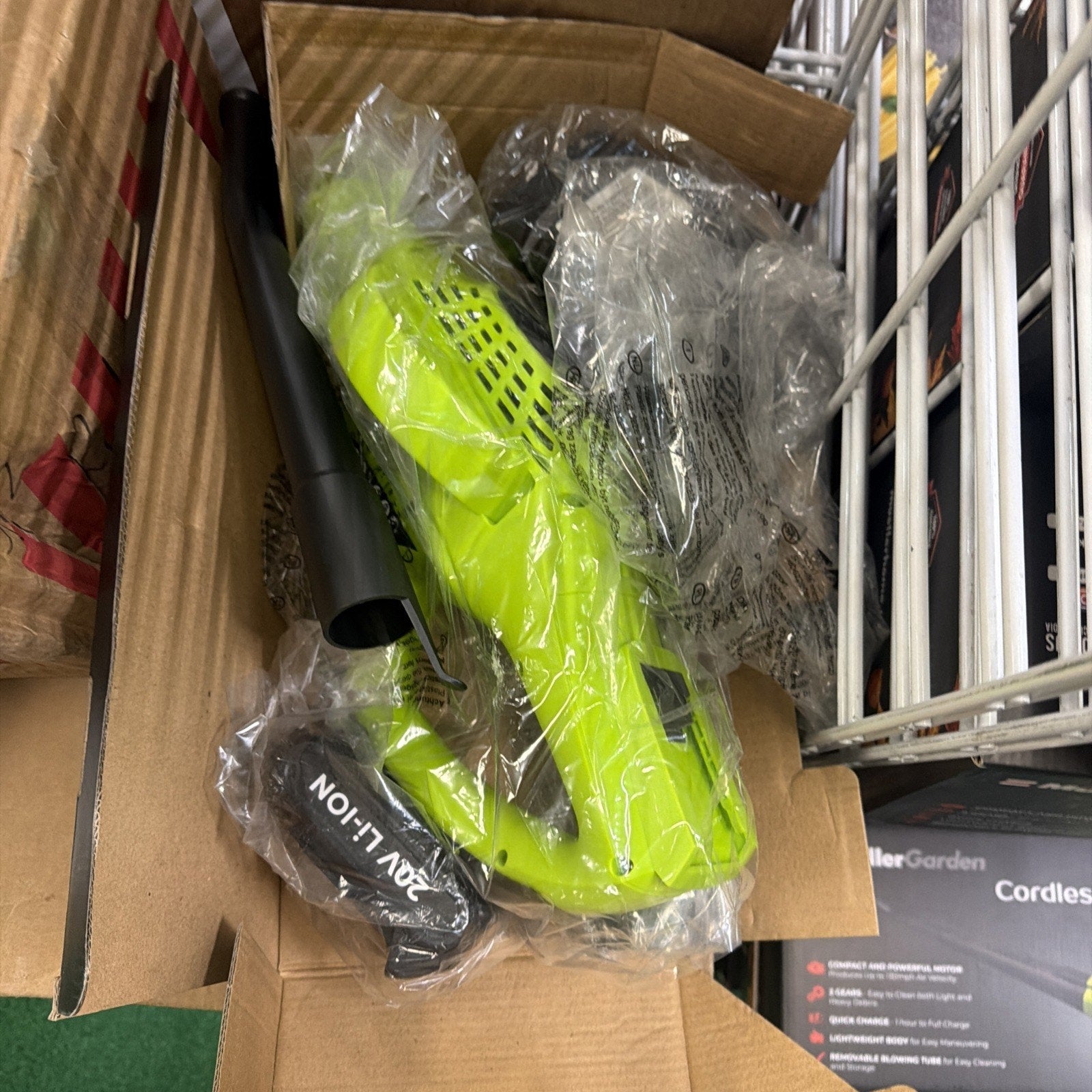 Bright green cordless handheld leaf blower with black detachable blower tube and 20V lithium battery, all wrapped in clear plastic inside a cardboard box.