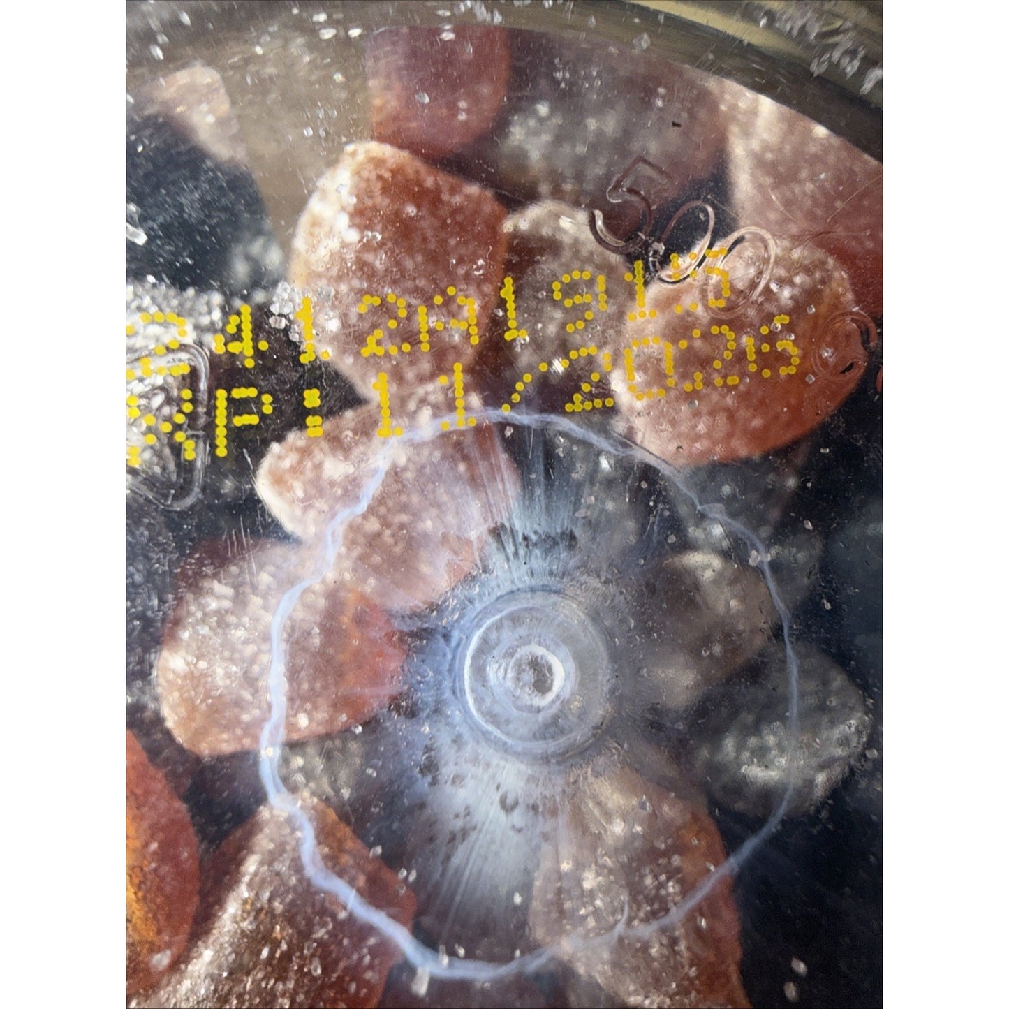 Close-up of a clear plastic container lid showing a printed expiration date with yellow text, with brown sugar-coated gummy candies visible inside the sealed container.