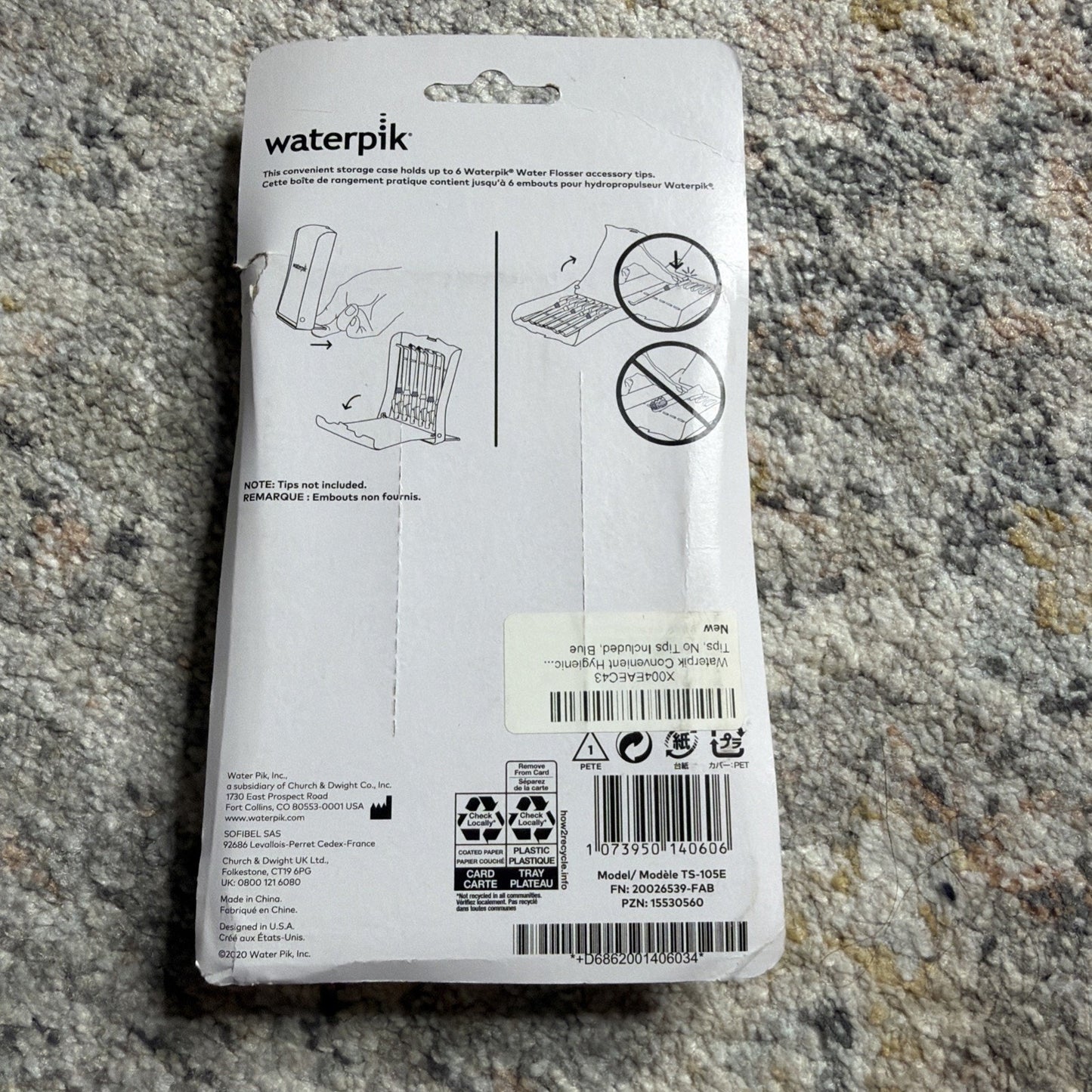 Back of Waterpik replacement tip storage case packaging showing instructions for holding up to eight Waterpik flosser tips with illustrations, text, and product codes on a gray carpet background.