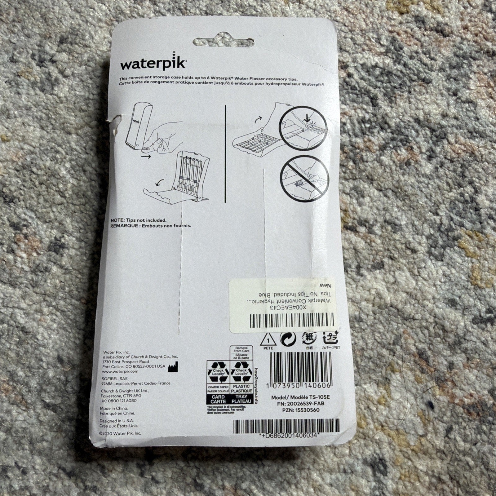 Back of Waterpik replacement tip storage case packaging showing instructions for holding up to eight Waterpik flosser tips with illustrations, text, and product codes on a gray carpet background.