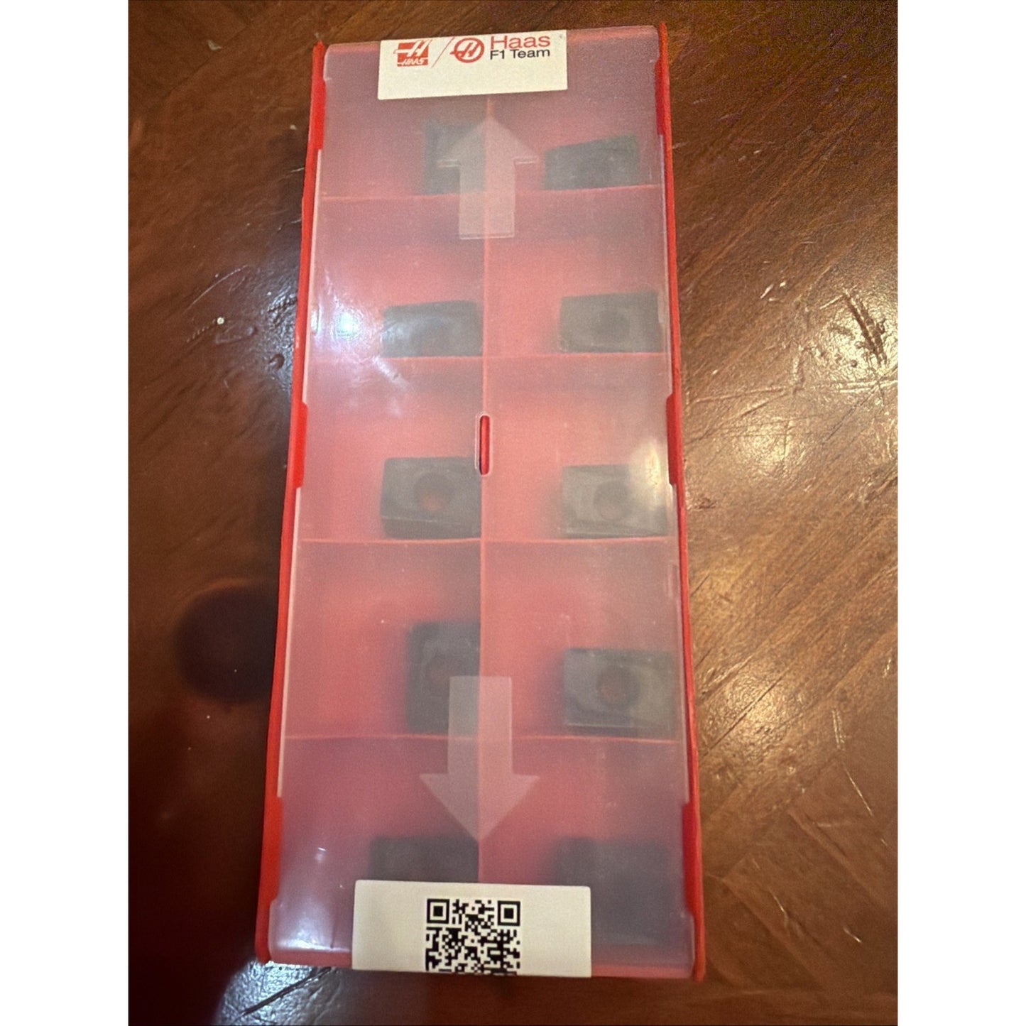 Rectangular red plastic storage box with a translucent lid containing eight small black inserts arranged in compartments, placed on a wooden surface with visible grain.