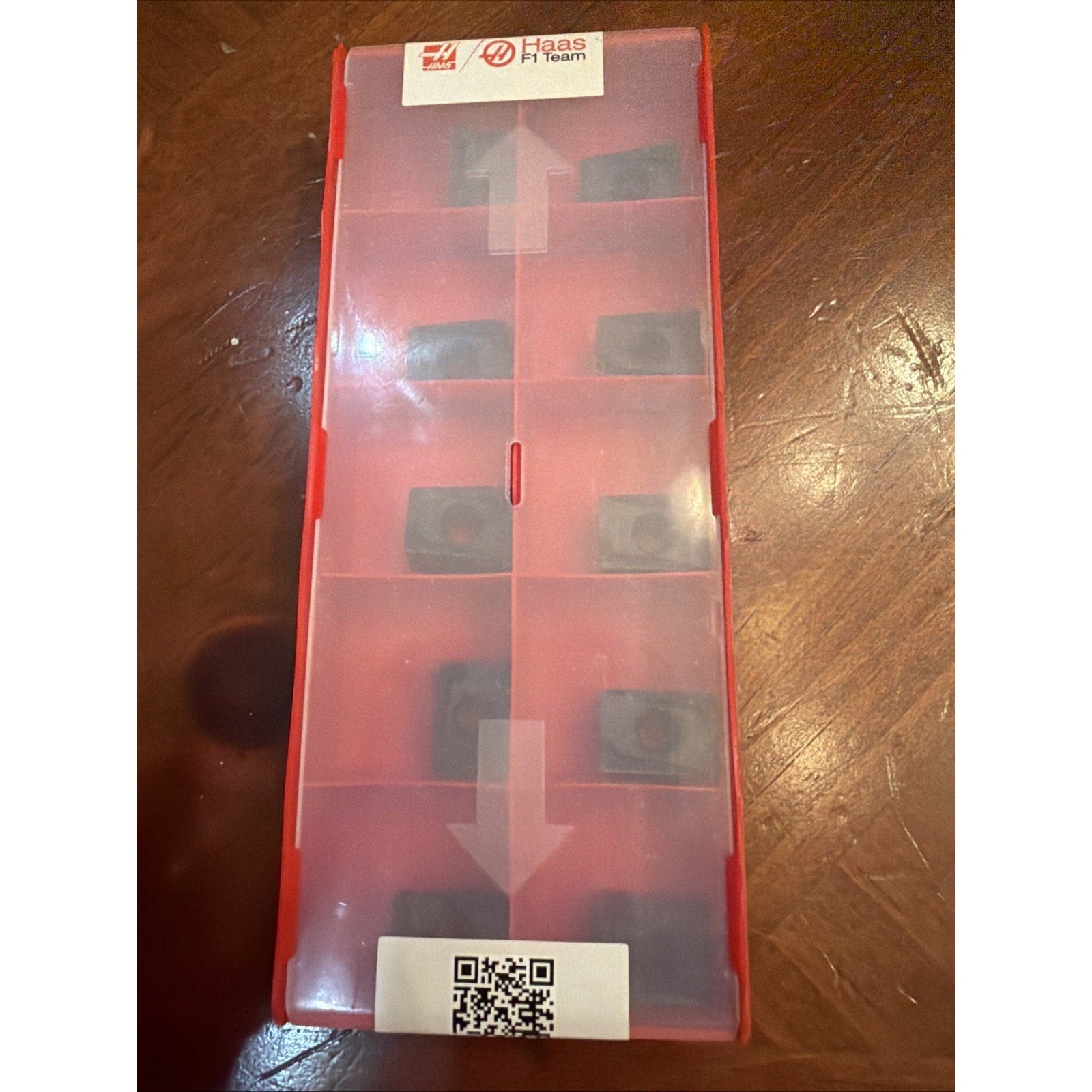Rectangular red plastic storage box with a translucent lid containing eight small black inserts arranged in compartments, placed on a wooden surface with visible grain.