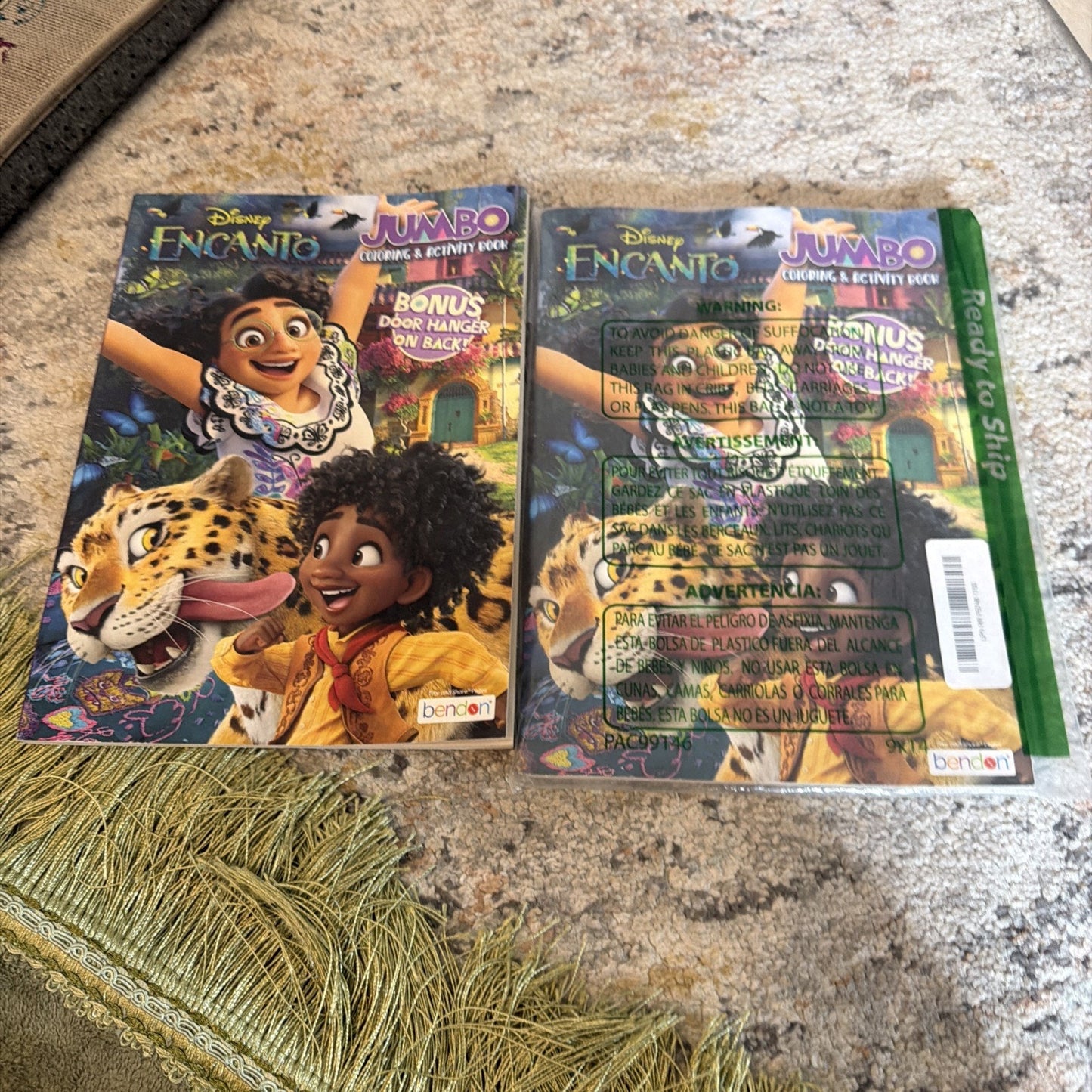 Two Disney Encanto-themed jumbo coloring and activity books side by side on a textured carpet, featuring Mirabel, Antonio, and a jaguar on colorful covers with bonus door hanger text.