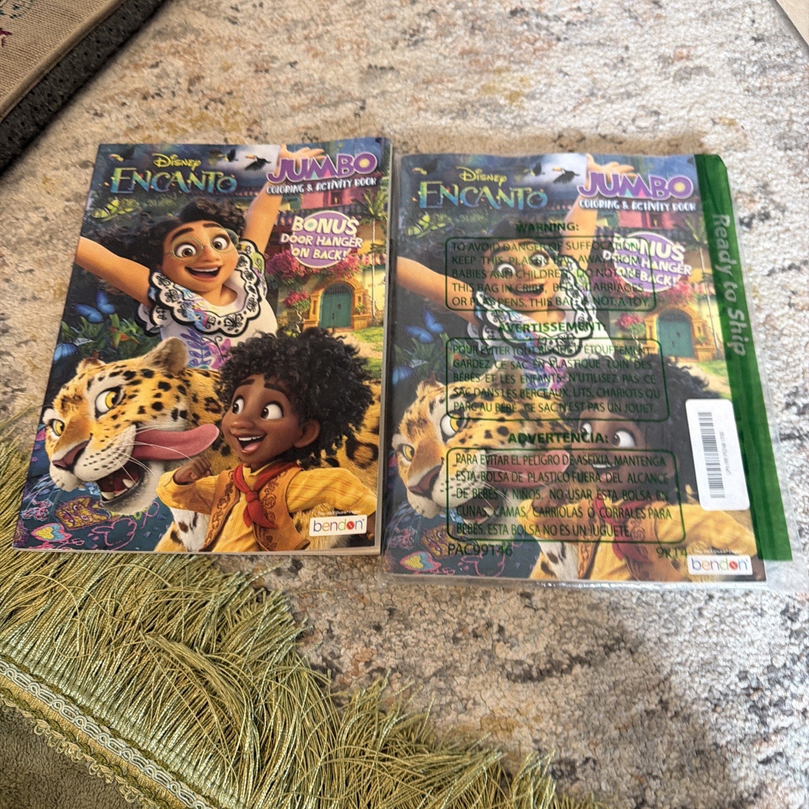 Two Disney Encanto-themed jumbo coloring and activity books side by side on a textured carpet, featuring Mirabel, Antonio, and a jaguar on colorful covers with bonus door hanger text.