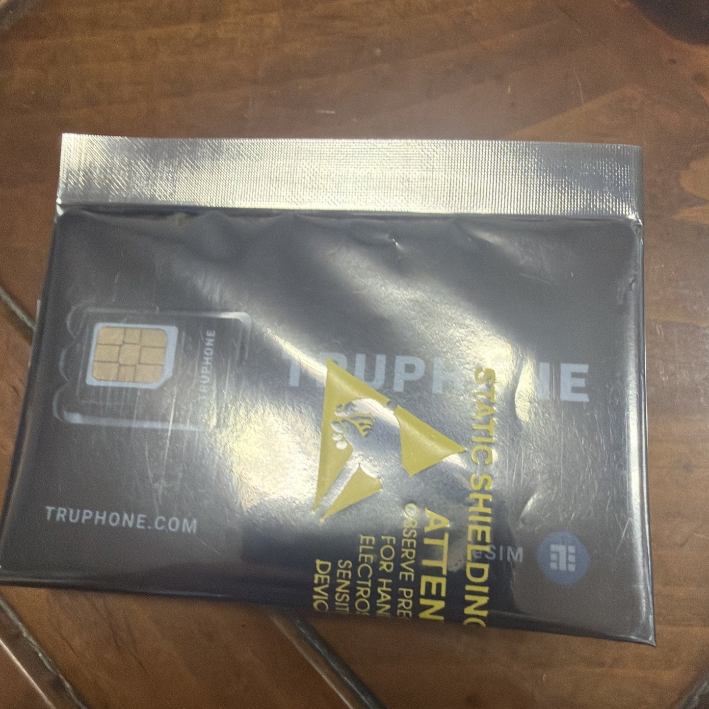 Black anti-static shielding bag containing a SIM card packet labeled with warnings to keep away from phone, electronic devices, and sensitive demos, placed on a wooden surface.