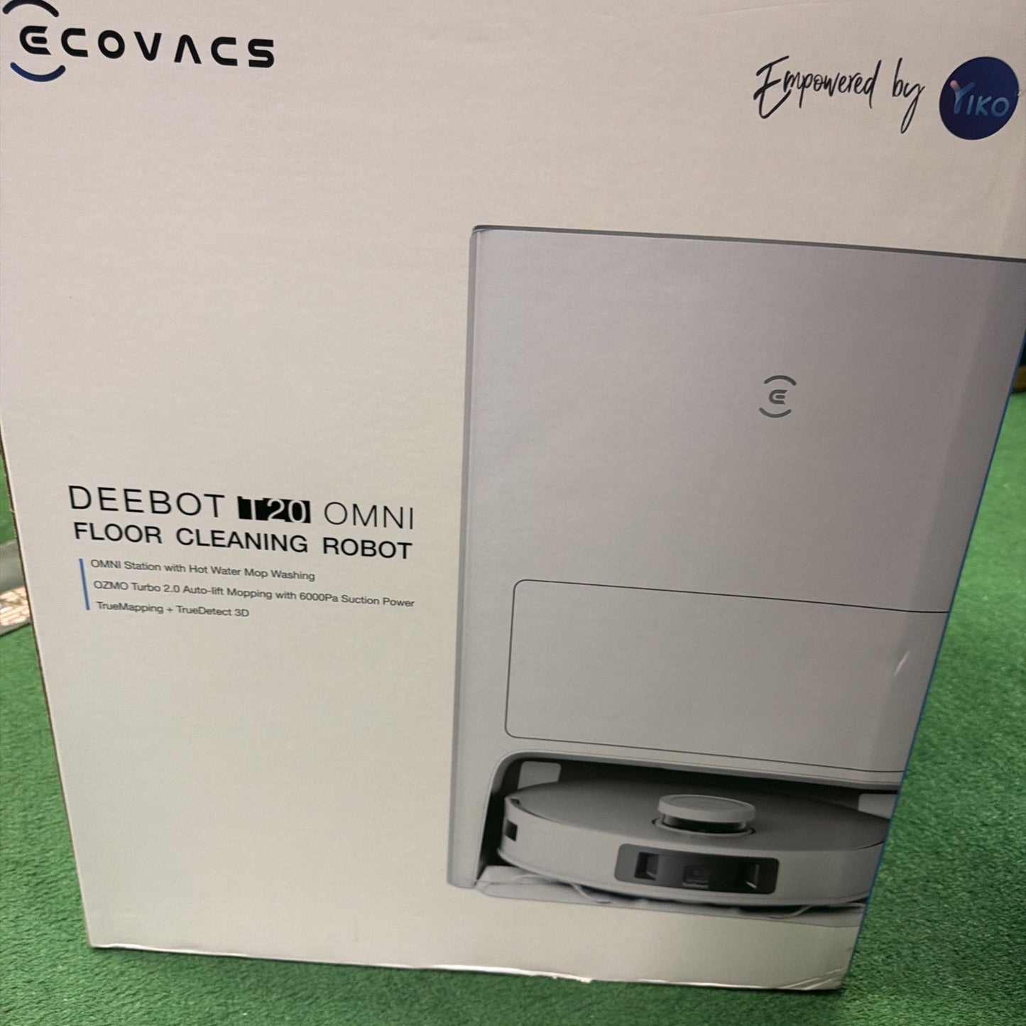 Packaging box showing a white floor cleaning robot partially visible from its storage station, featuring branding and product details on a white background with a green carpet underneath.
