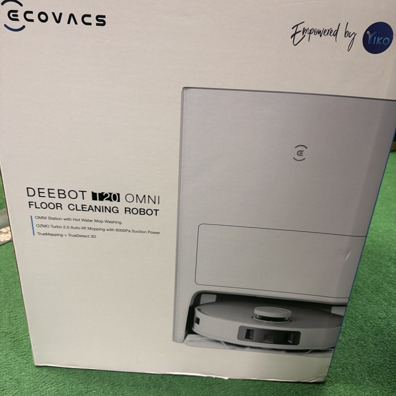 Packaging box showing a white floor cleaning robot partially visible from its storage station, featuring branding and product details on a white background with a green carpet underneath.