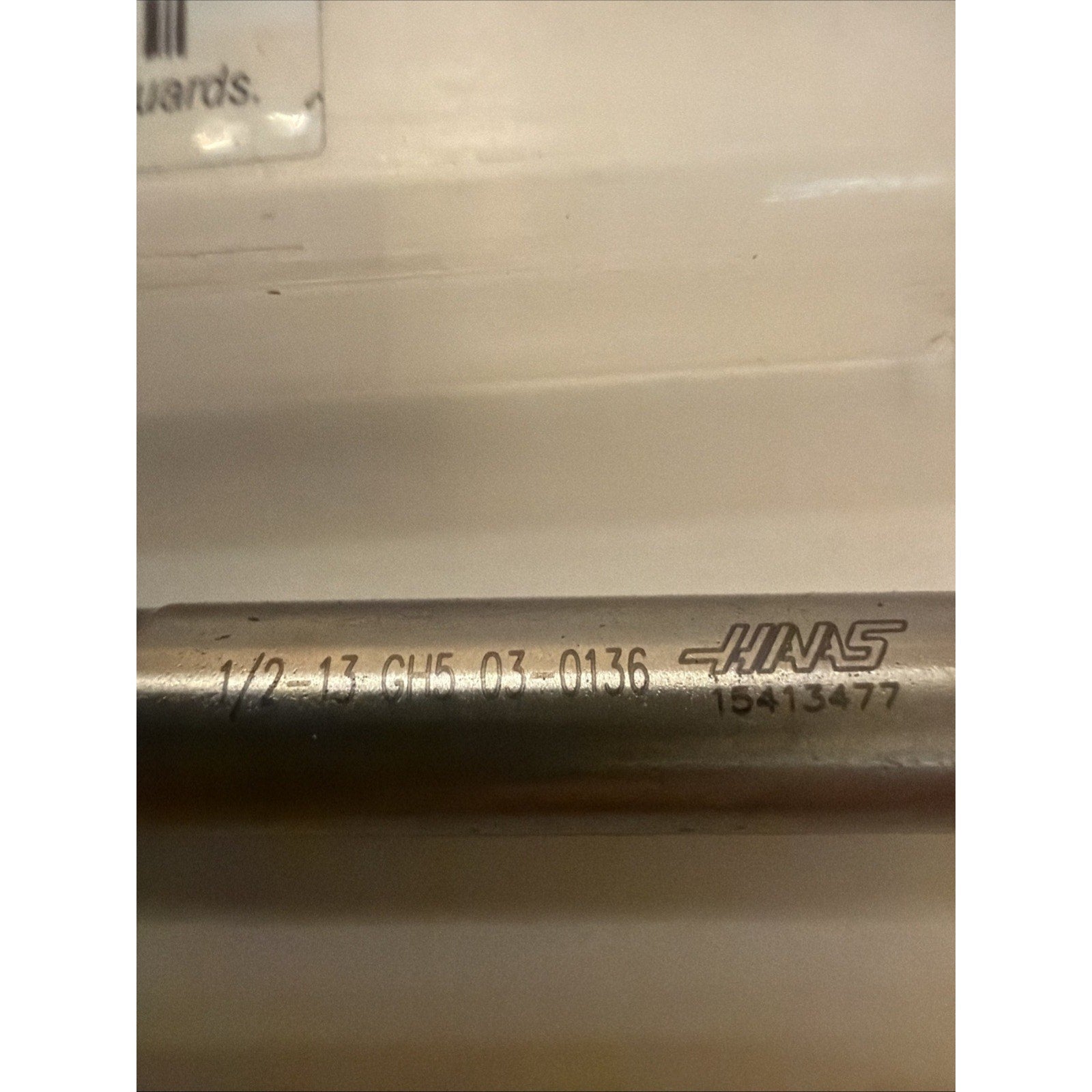 Close-up of a cylindrical metal tool with engraved alphanumeric markings and a Haas logo, placed on a white surface under soft lighting.