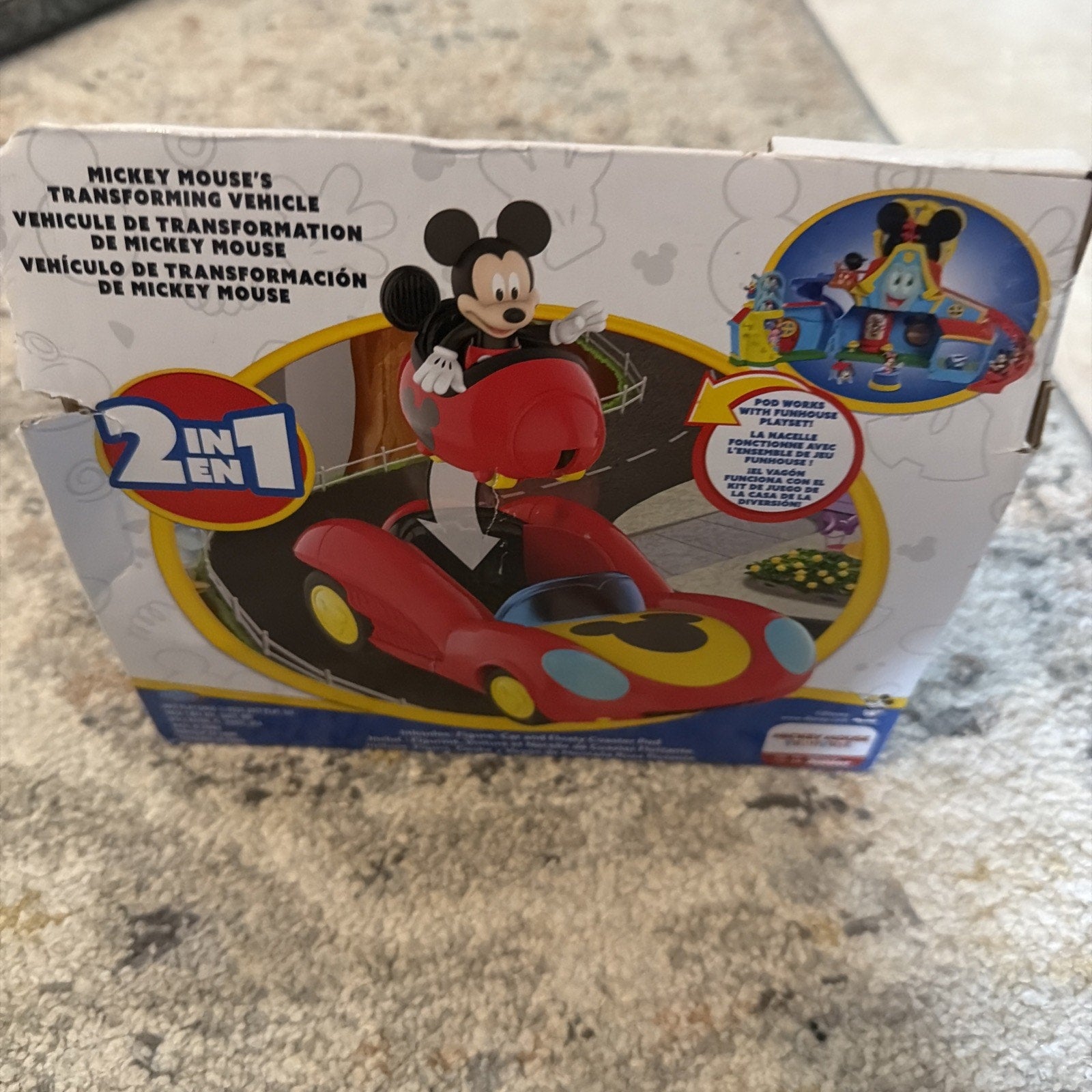 Box of a 2-in-1 Mickey Mouse transforming vehicle toy featuring Mickey sitting in a red car that converts into a playhouse, displayed on a carpeted surface.