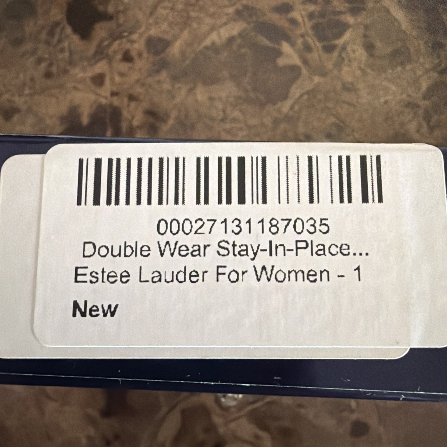 Close-up of a white product label with a barcode and text reading "Double Wear Stay-In-Place... Estee Lauder For Women - 1 New" on a dark blue box against a textured brown surface.