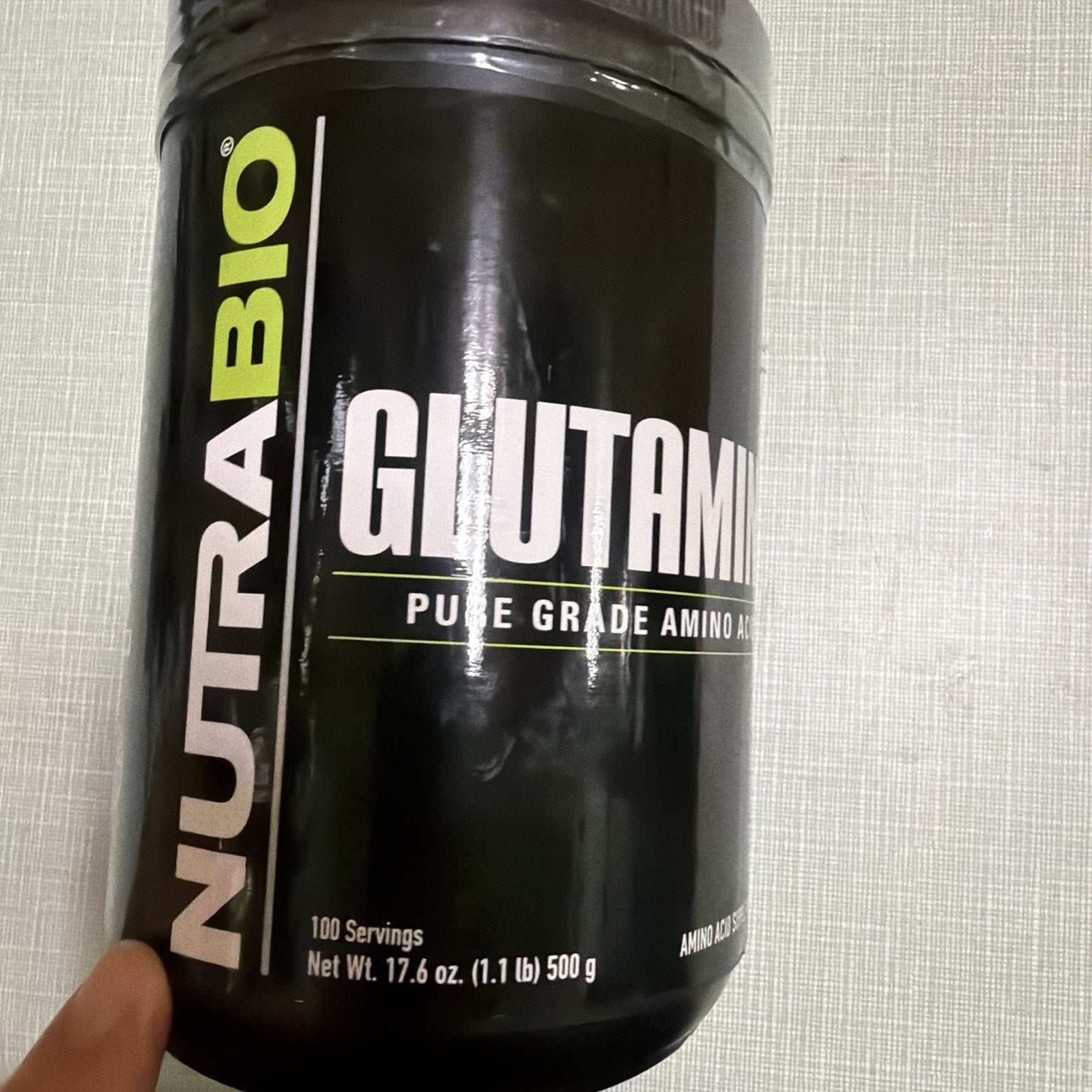 Black container of NutraBio Glutamine supplement labeled as pure grade amino acid with 100 servings and net weight of 500 grams held against a light textured background.