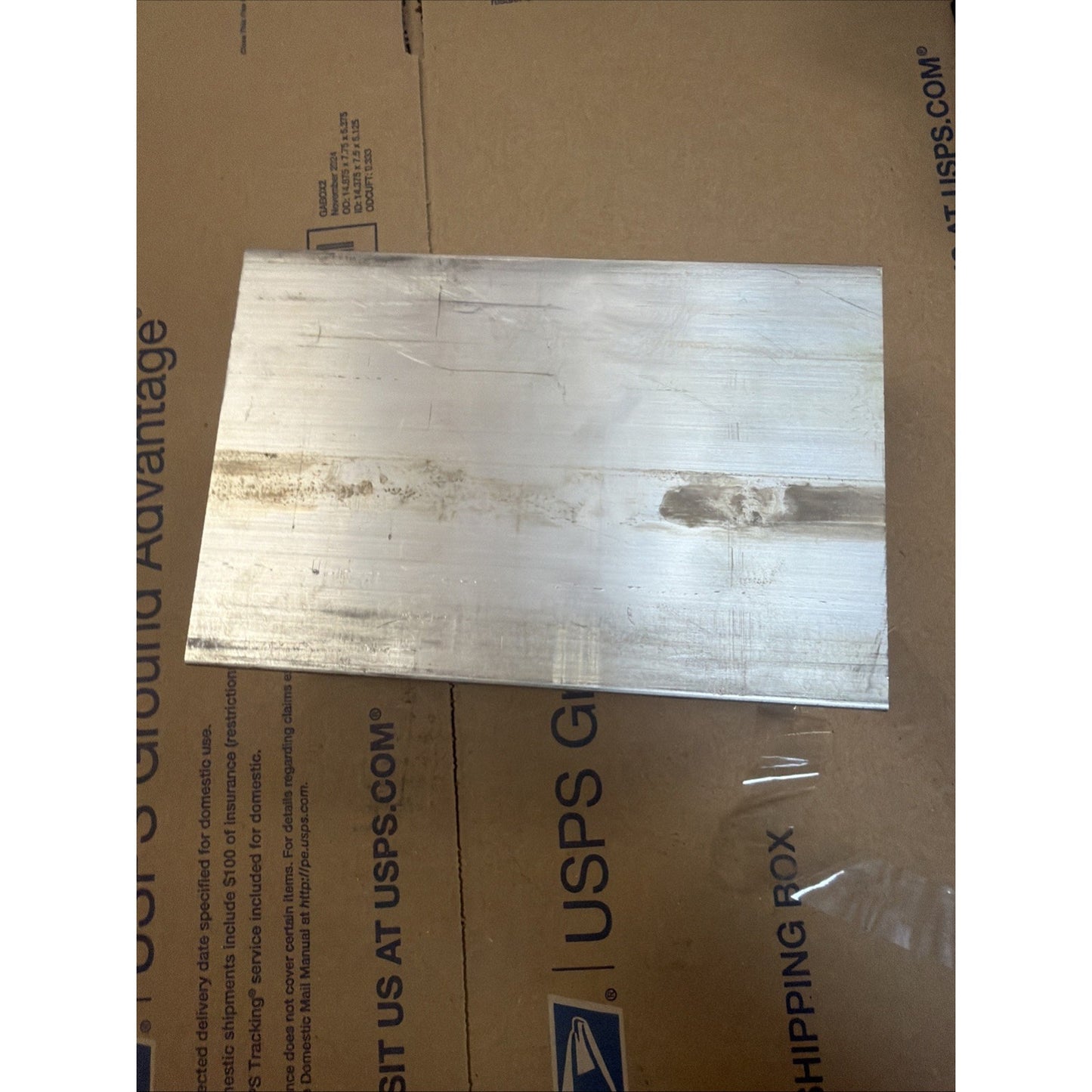 Rectangular metallic block with a scratched and slightly tarnished surface, resting on a USPS-branded cardboard shipping box with some clear plastic tape nearby.