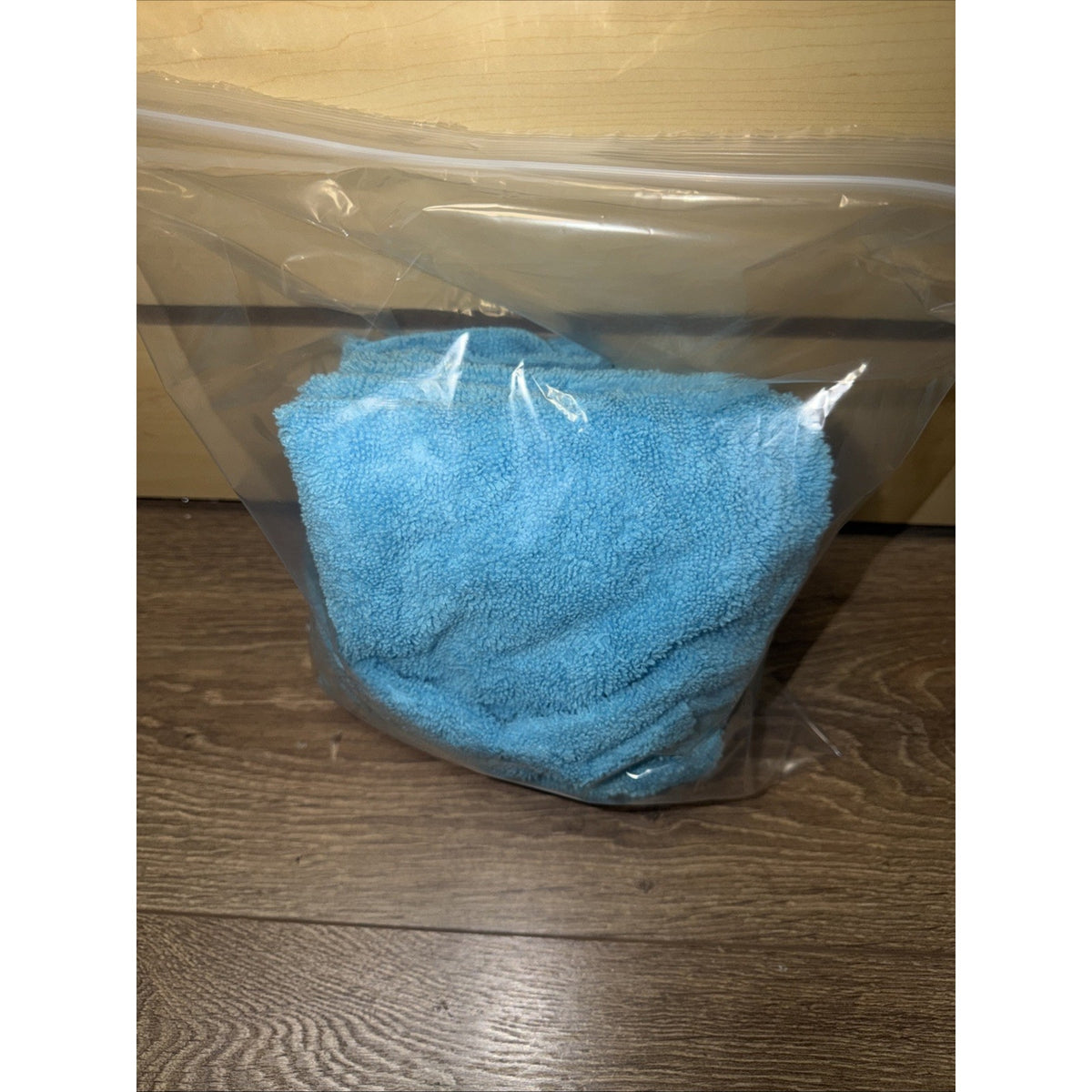 Light blue microfiber cloth folded inside a clear resealable plastic bag placed on a wooden surface with a light wooden background.