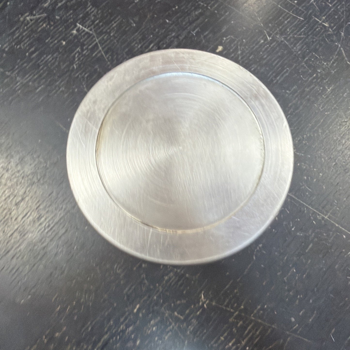 Brushed metal circular object with a recessed inner circle, resting on a scratched dark surface showing fine circular machining marks.