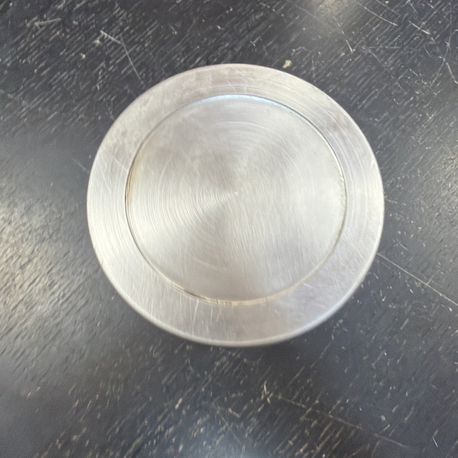 Brushed metal circular object with a recessed inner circle, resting on a scratched dark surface showing fine circular machining marks.