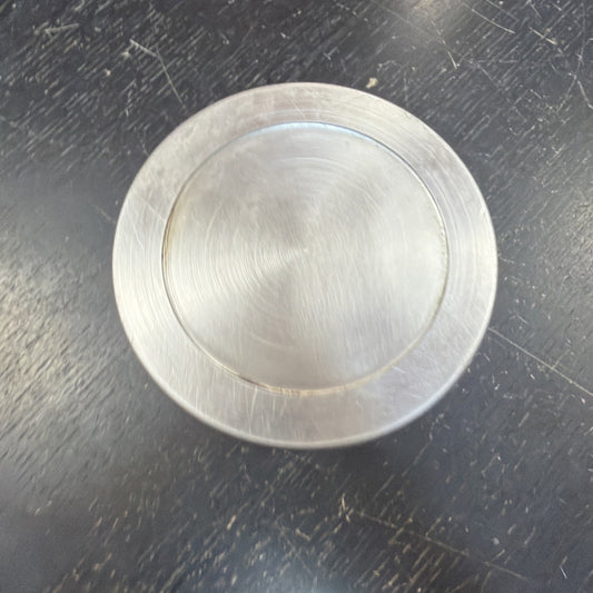 Brushed metal circular object with a recessed inner circle, resting on a scratched dark surface showing fine circular machining marks.