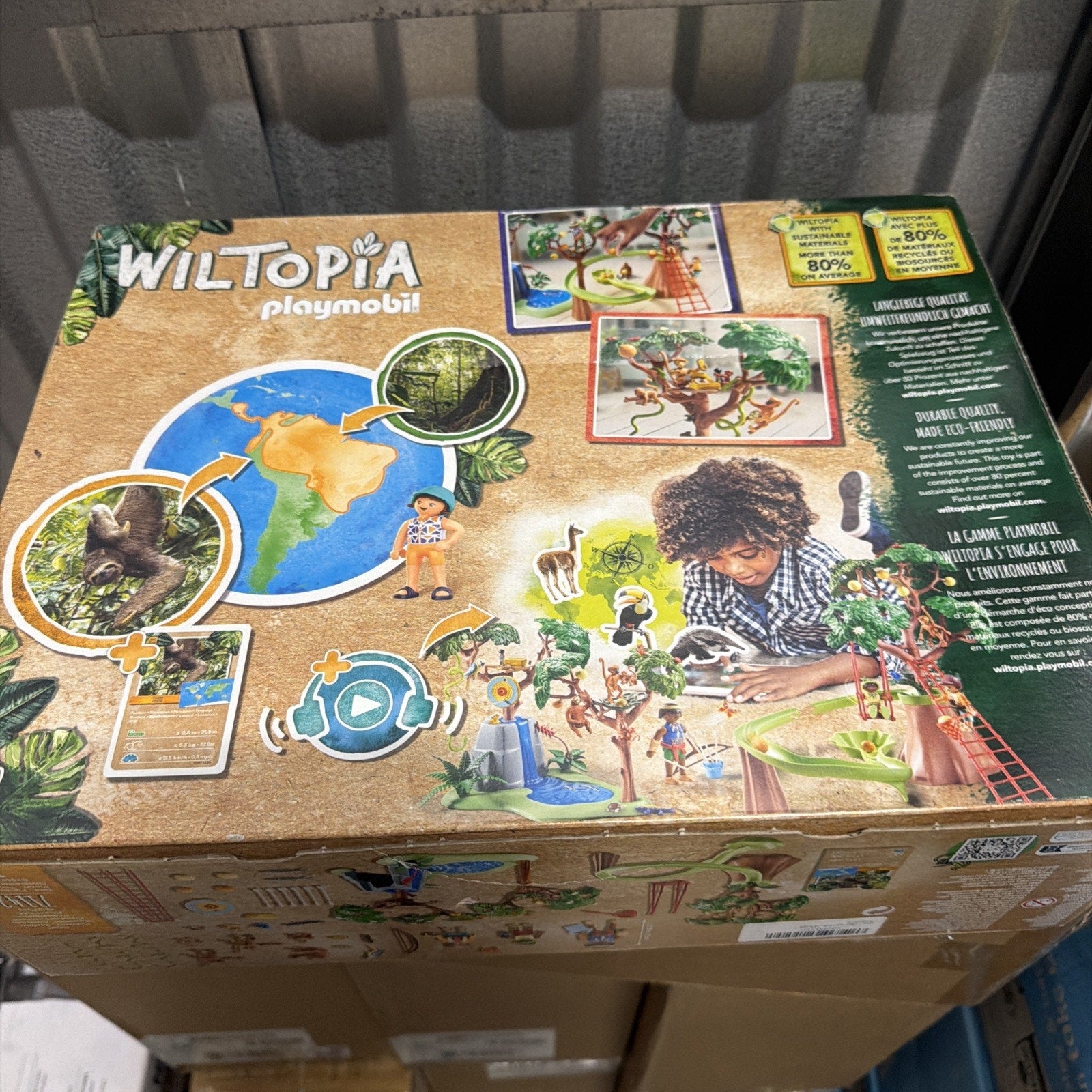 Top view of a Wiltopia Playmobil set box featuring illustrations of wildlife, a child playing with animal and forest figures, a world map with highlighted rainforest area, and text highlighting environmental education and sustainability features.