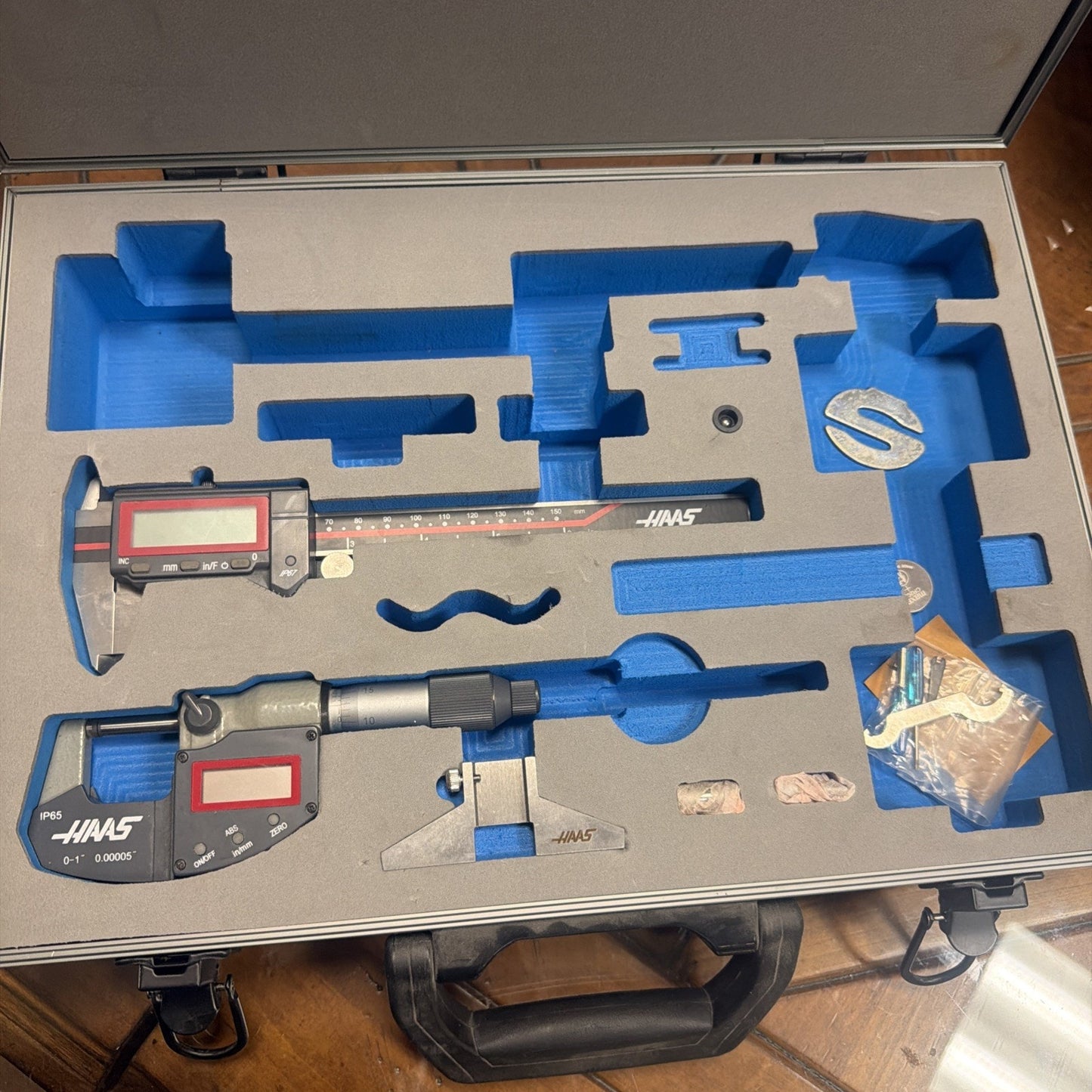 Open carrying case with custom gray foam inserts holding two digital measuring tools, a small metal circular part, and a plastic bag with additional accessories on a wooden floor.