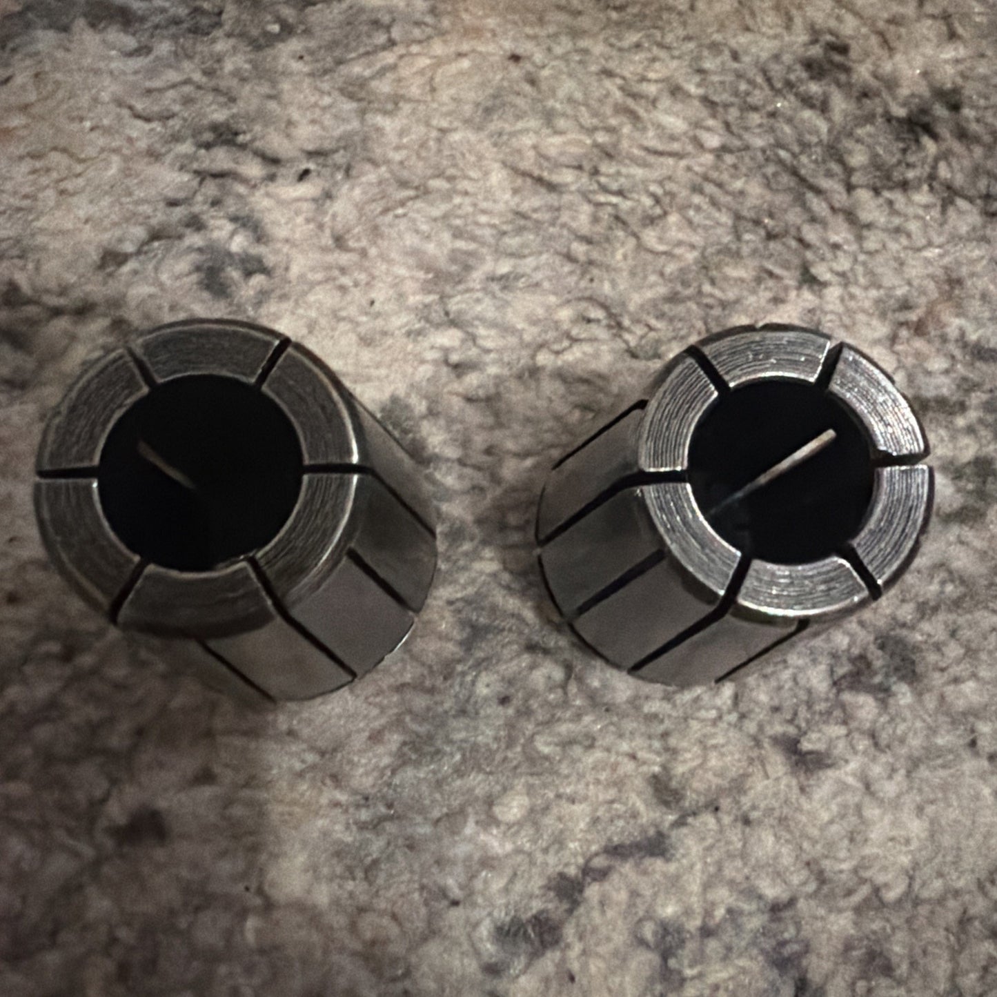 Two metallic cylindrical collets with multiple radial slits, placed upright on a textured carpet surface with a neutral color tone.