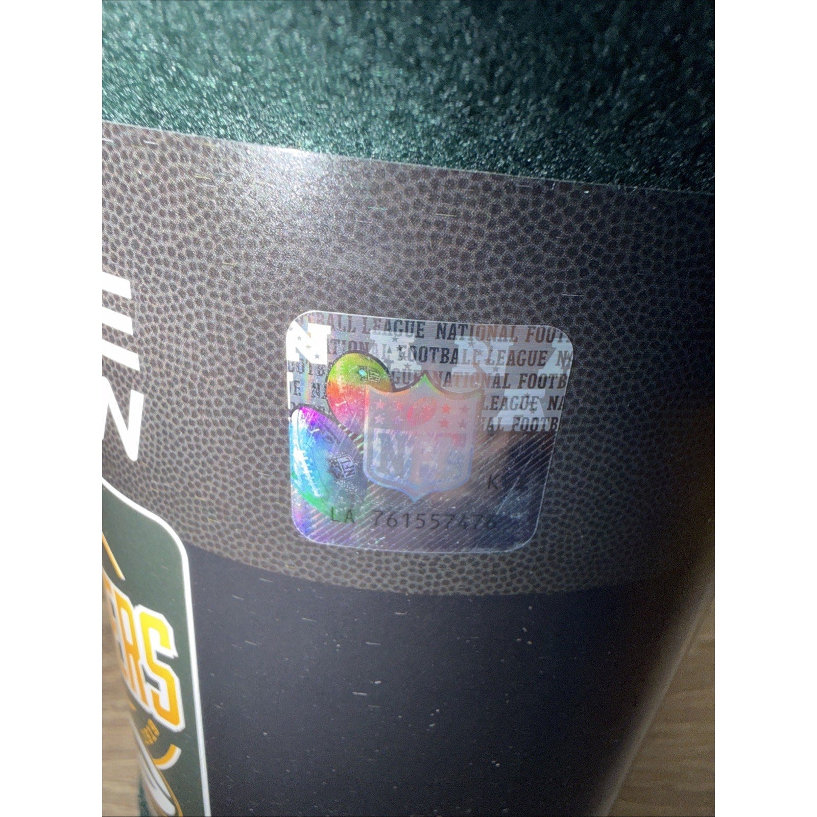 Close-up of a holographic NFL authenticity sticker with serial number on textured dark packaging featuring a brown pattern resembling a football surface.