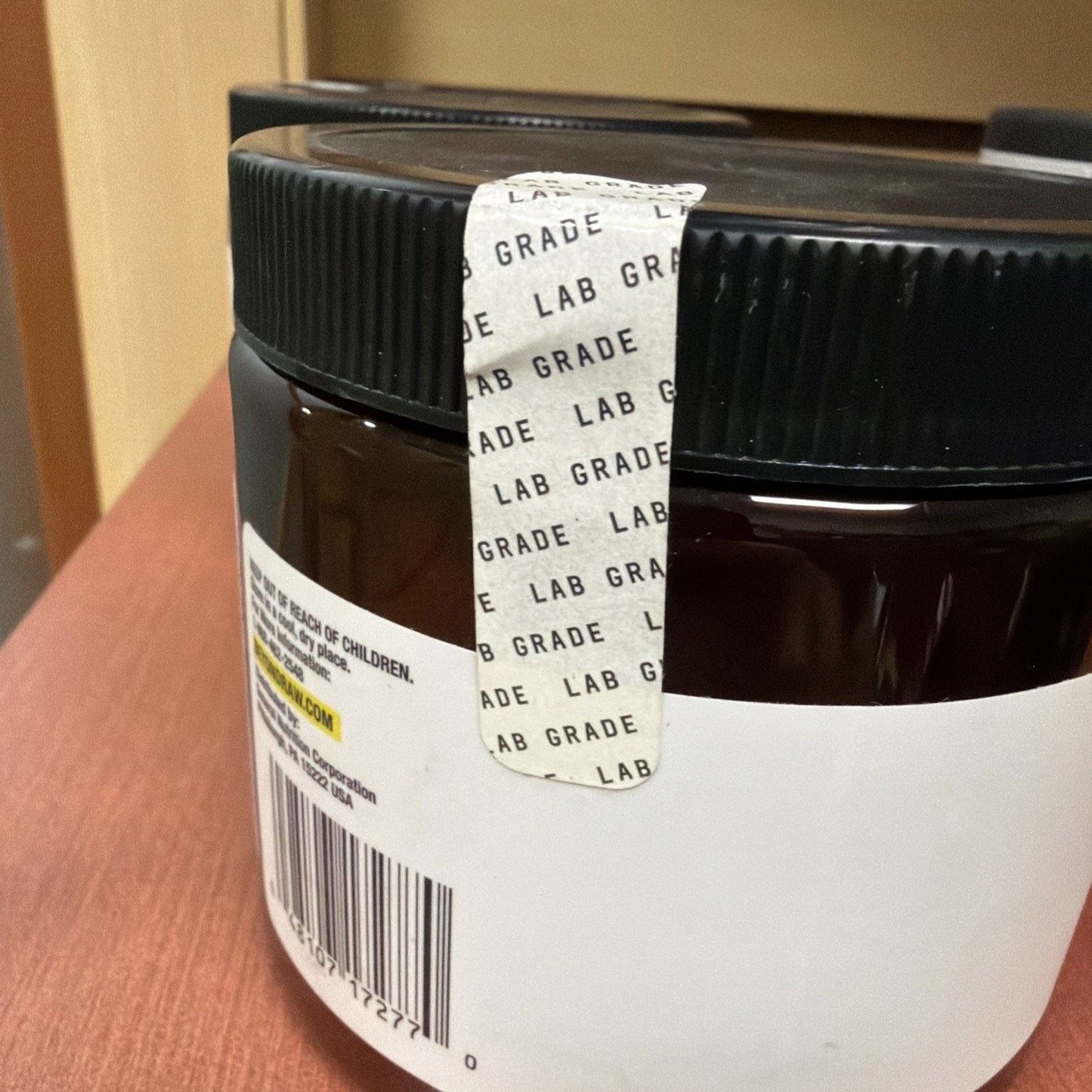Close-up of a dark amber plastic container with a black screw-on lid sealed with a white tamper-evident strip marked "LAB GRADE," resting on a wooden surface.