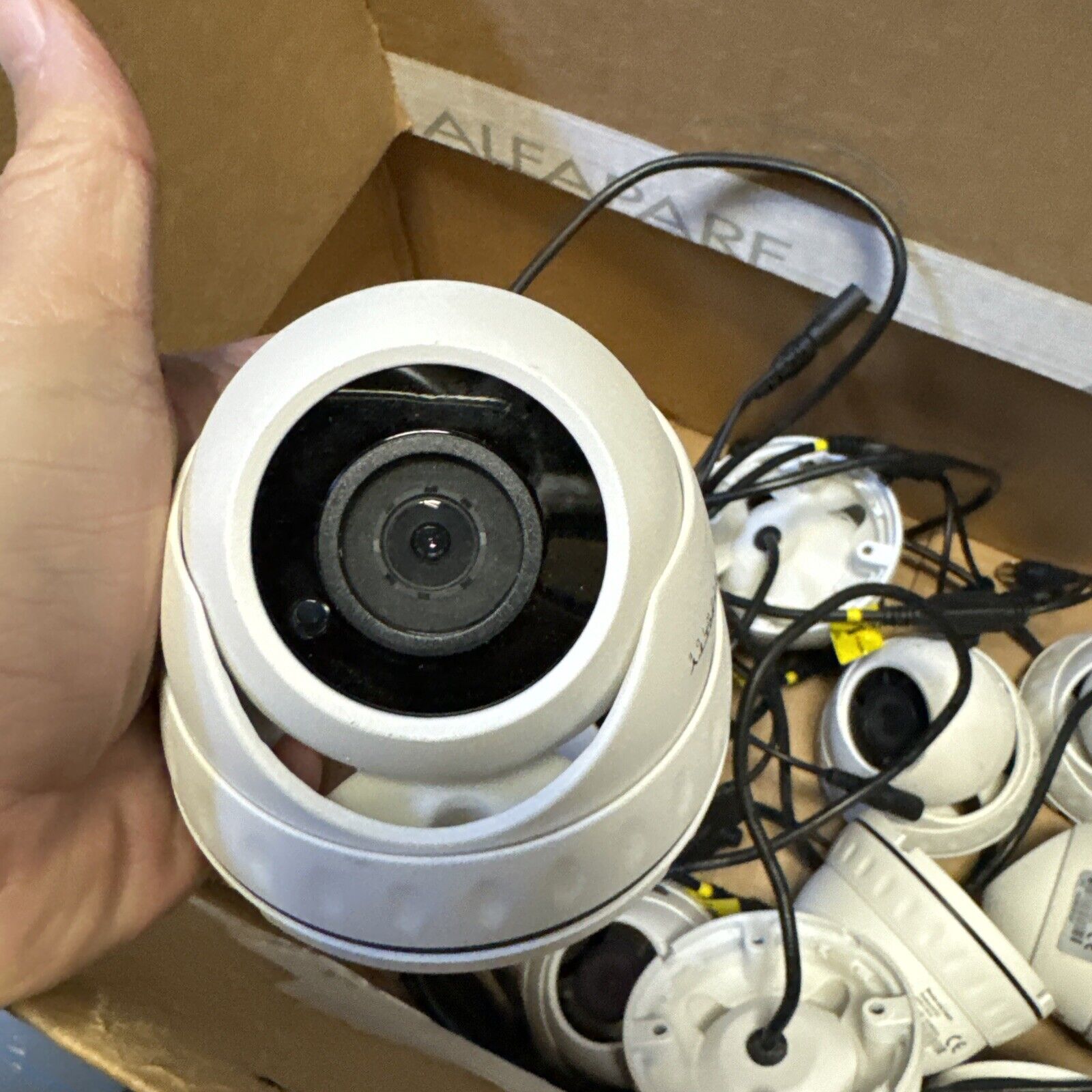 Hand holding a white Boss 1080P security camera with multiple wired cameras in a cardboard box background
