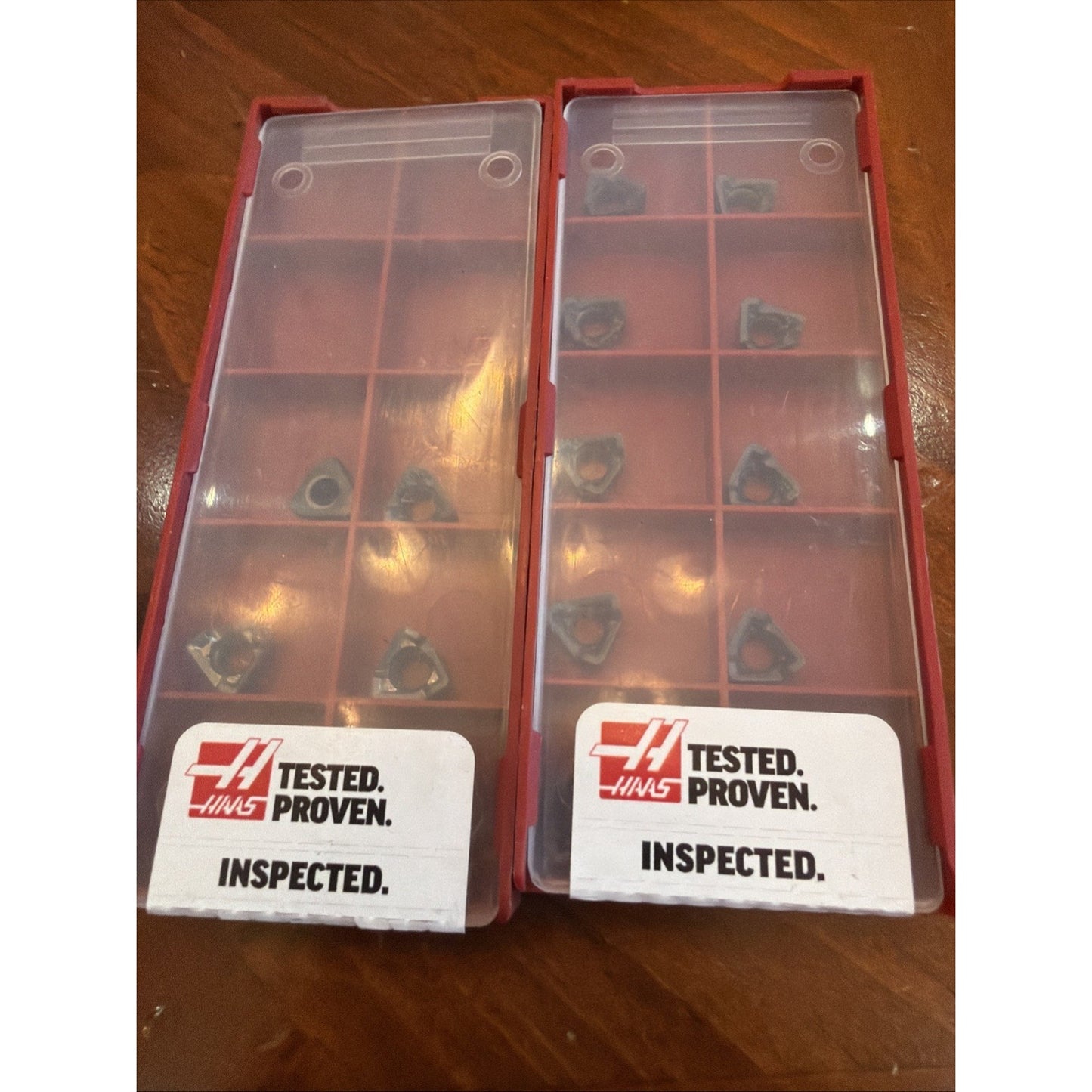 Two red plastic storage cases with transparent lids containing small triangular metal inserts, each labeled with "Haas Tested. Proven. Inspected." on a wooden surface.