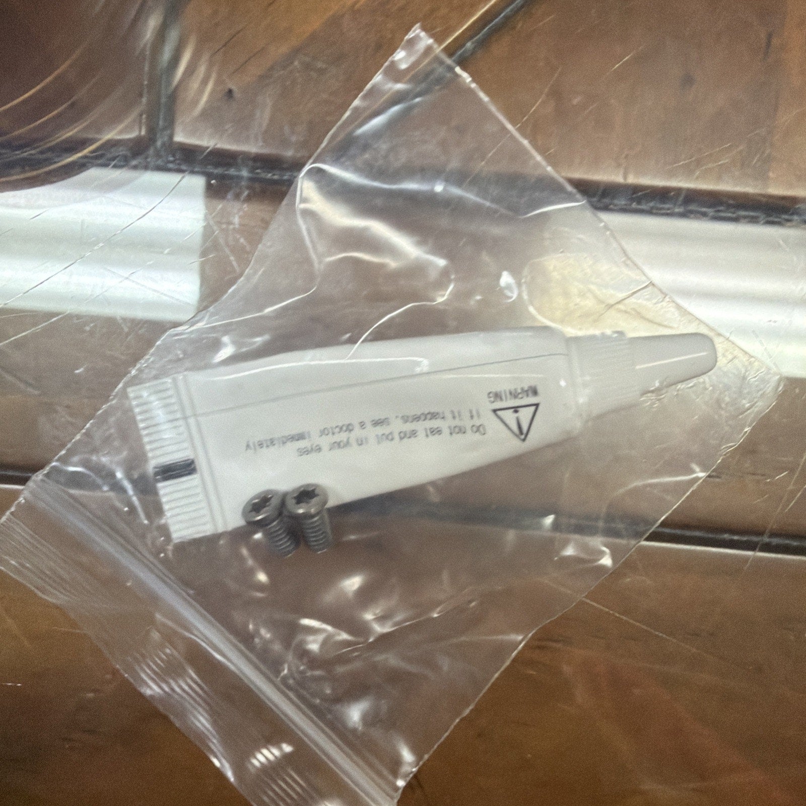 Small white plastic tube with printed warning text and three small silver screws, sealed inside a clear plastic bag on a reflective surface.