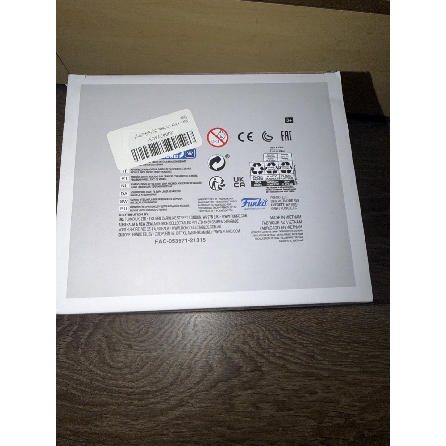 Back of a white product box featuring multiple certification logos, safety warnings, barcode sticker, multilingual text, and Funko branding on a wooden surface.
