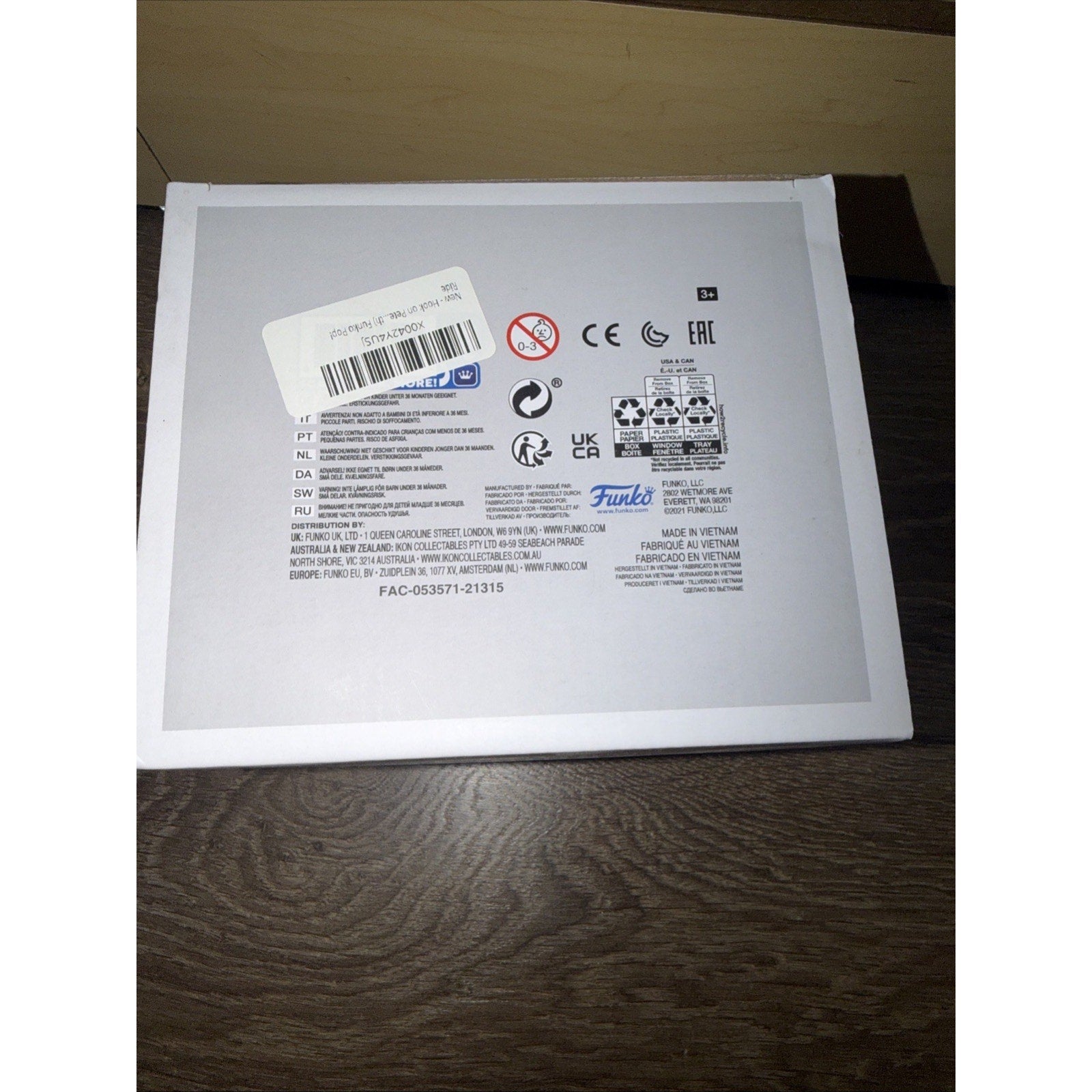 Back of a white product box featuring multiple certification logos, safety warnings, barcode sticker, multilingual text, and Funko branding on a wooden surface.