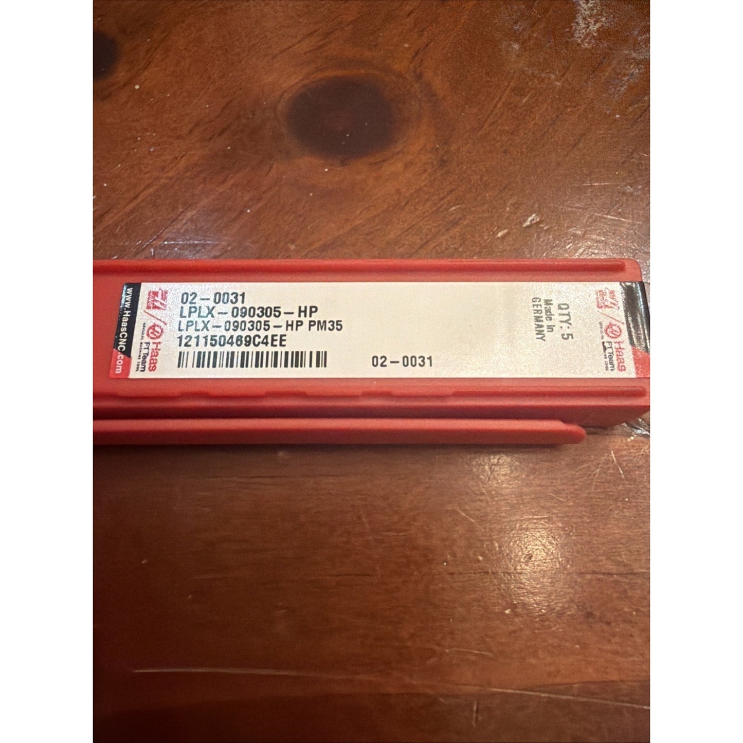 Red rectangular plastic case with a white label showing product code "LPLX-090305-HP PM35," a barcode, quantity "Qty. 15," and "Made in Germany," resting on a wooden surface.