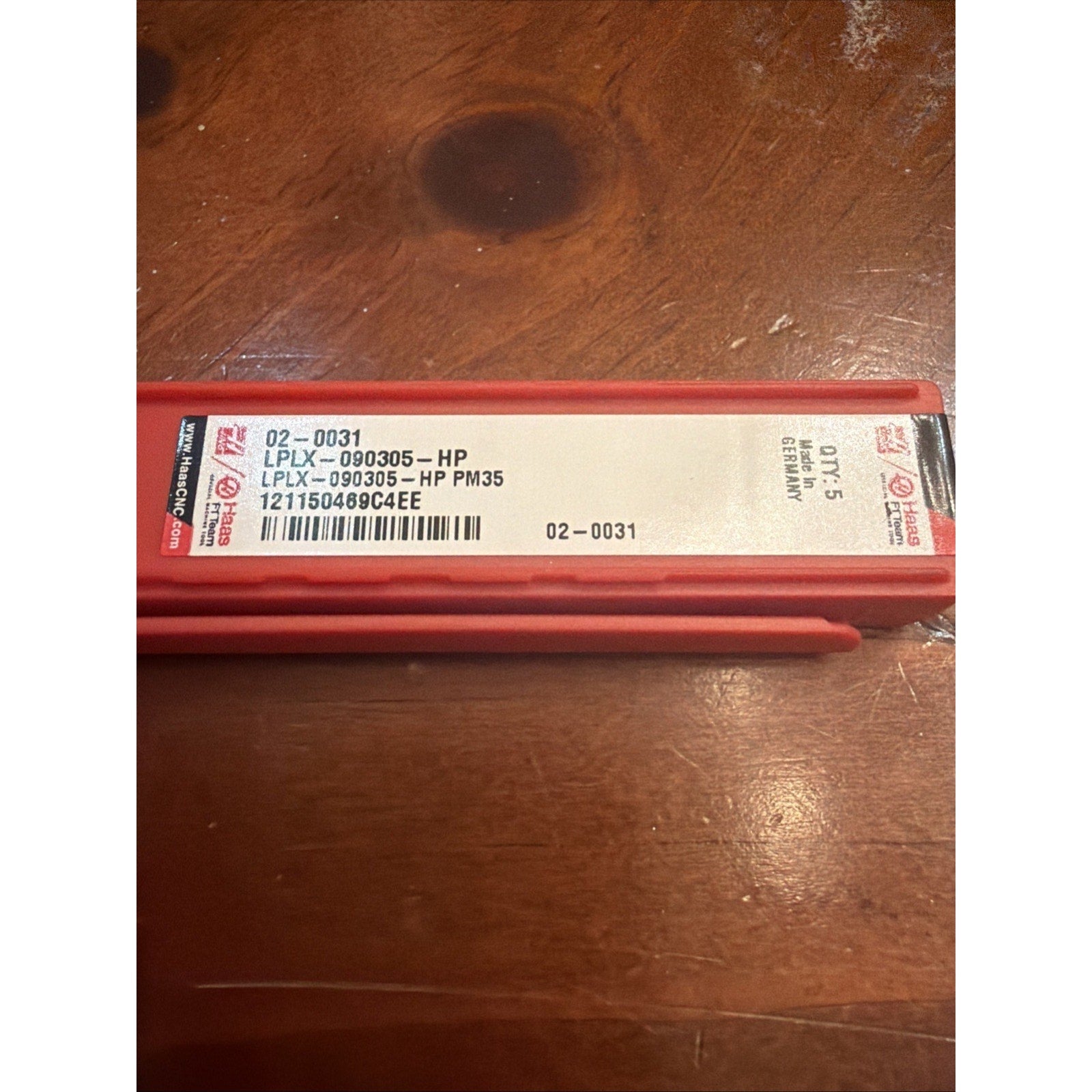 Red rectangular plastic case with a white label showing product code "LPLX-090305-HP PM35," a barcode, quantity "Qty. 15," and "Made in Germany," resting on a wooden surface.