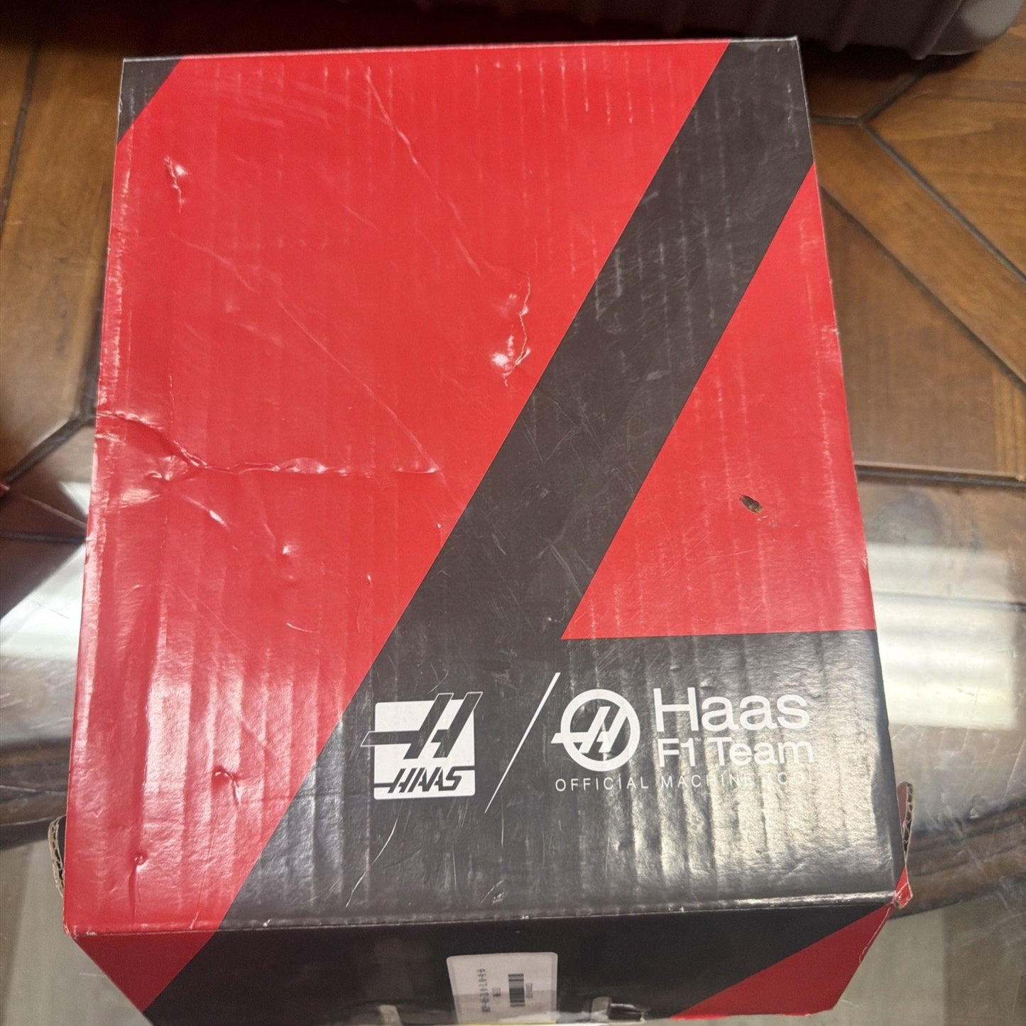 Red and black cardboard box with Haas and Haas F1 Team logos printed on the top, placed on a wooden surface reflecting light.