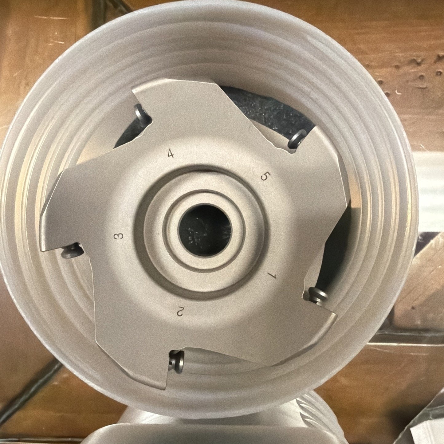 Top-down view of a circular metal cutting tool with five numbered teeth, mounted inside a white plastic cylindrical holder on a wooden surface.